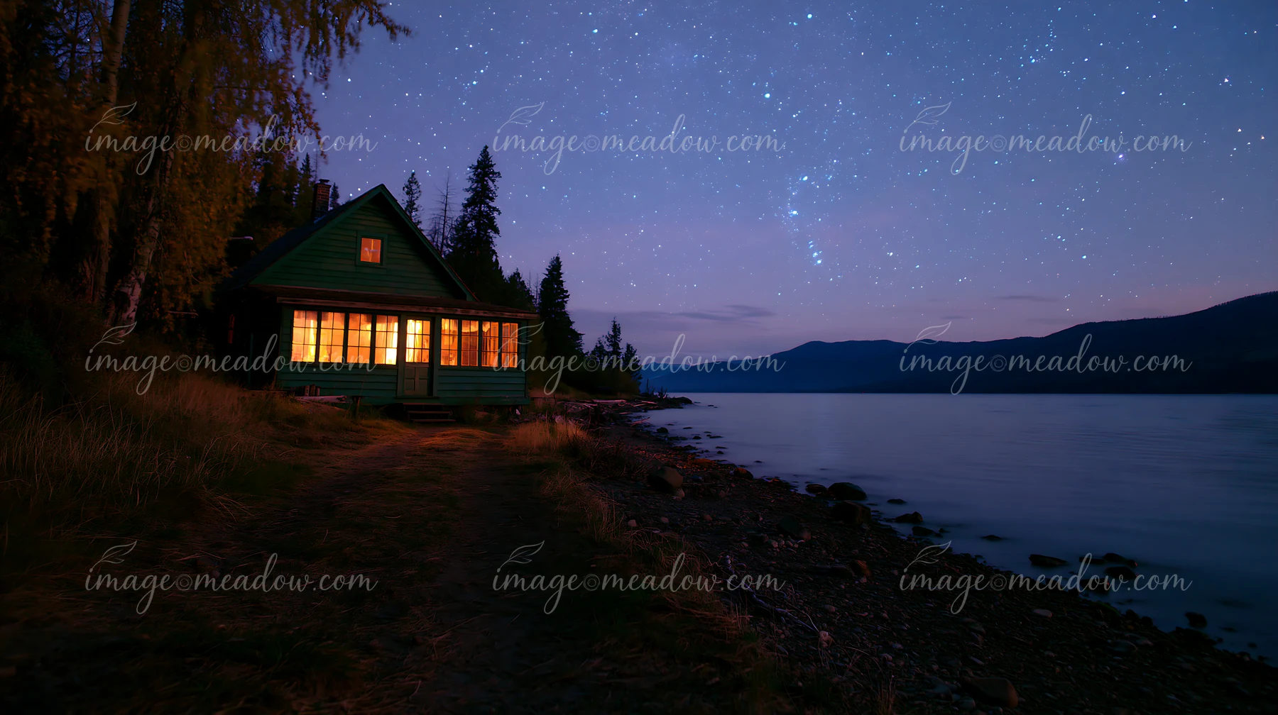 A cabin stands illuminated by warm light under a starry sky at night near a calm lake.