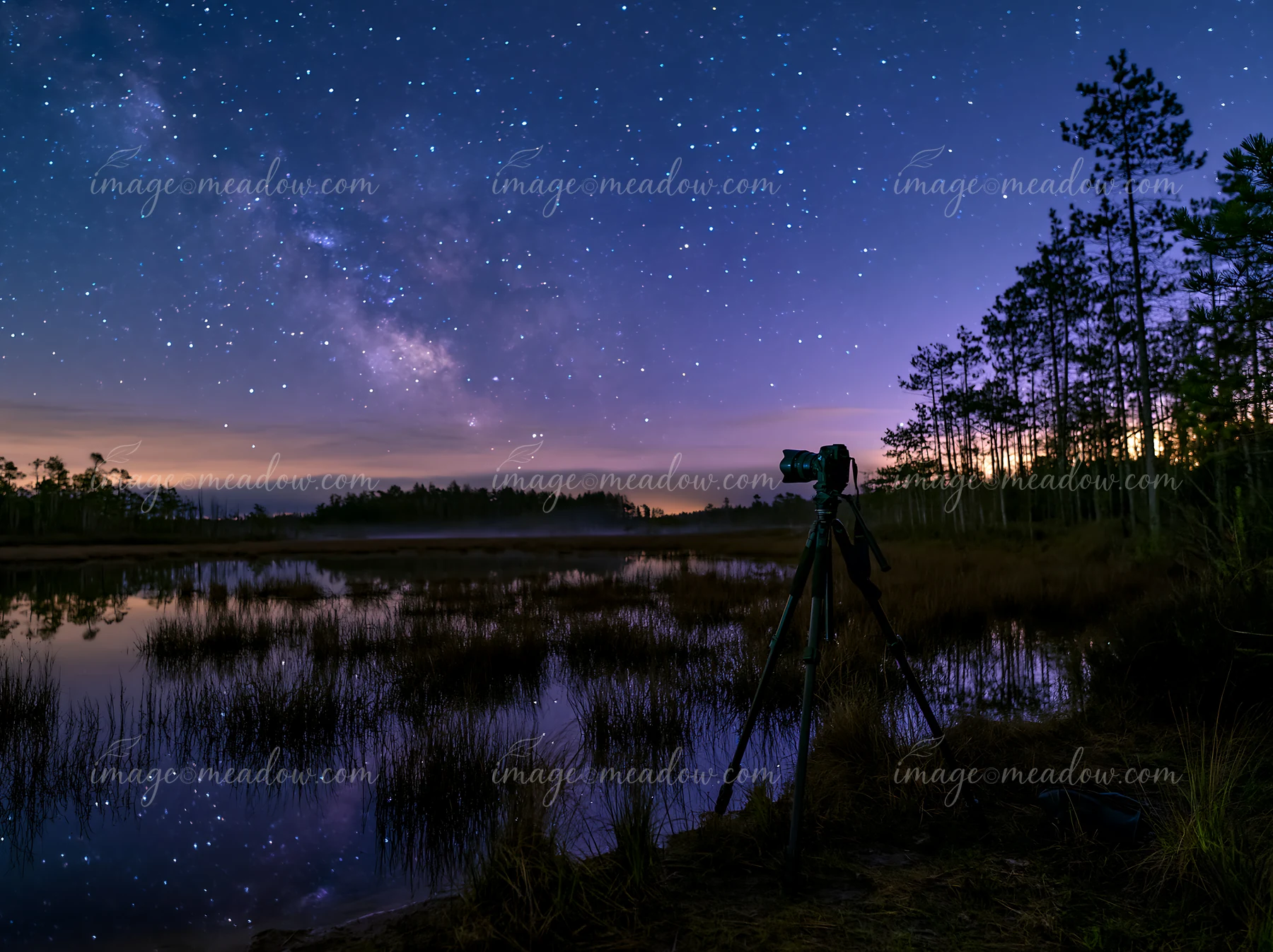 A camera tripod stands by a calm lake reflecting stars and the Milky Way.