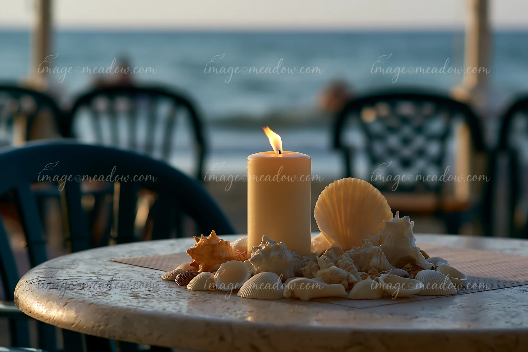 A candle surrounded by seashells sits on a table near the beach.