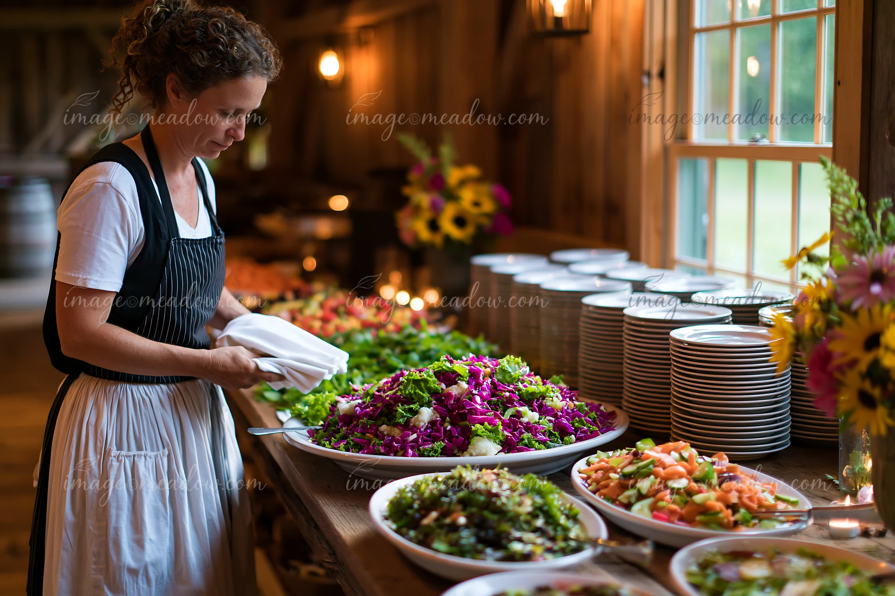 A chef arranges a variety of fresh salads at a banquet table in a cozy barn.