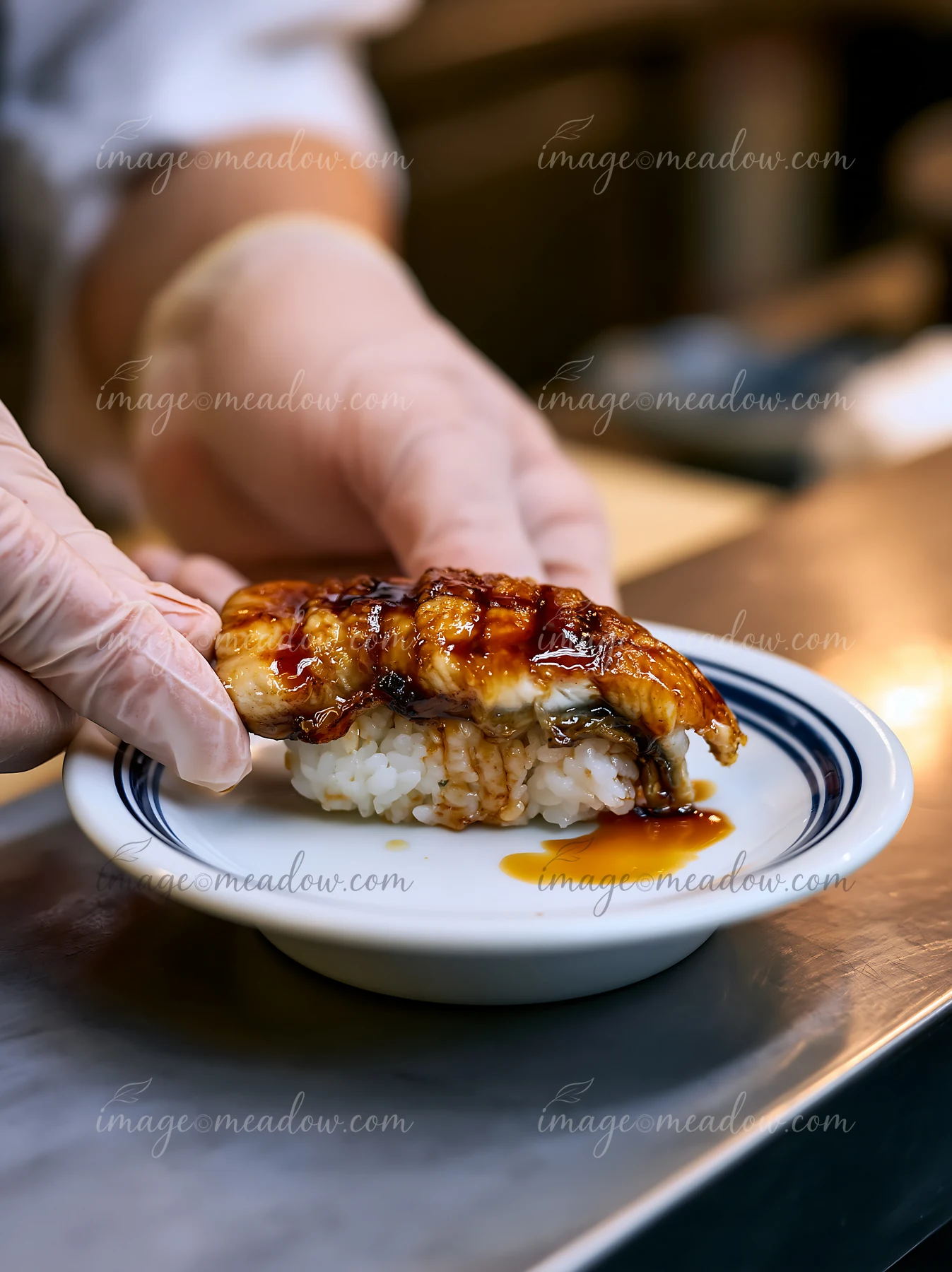 A chef serves a single eel nigiri with sauce on a small plate.