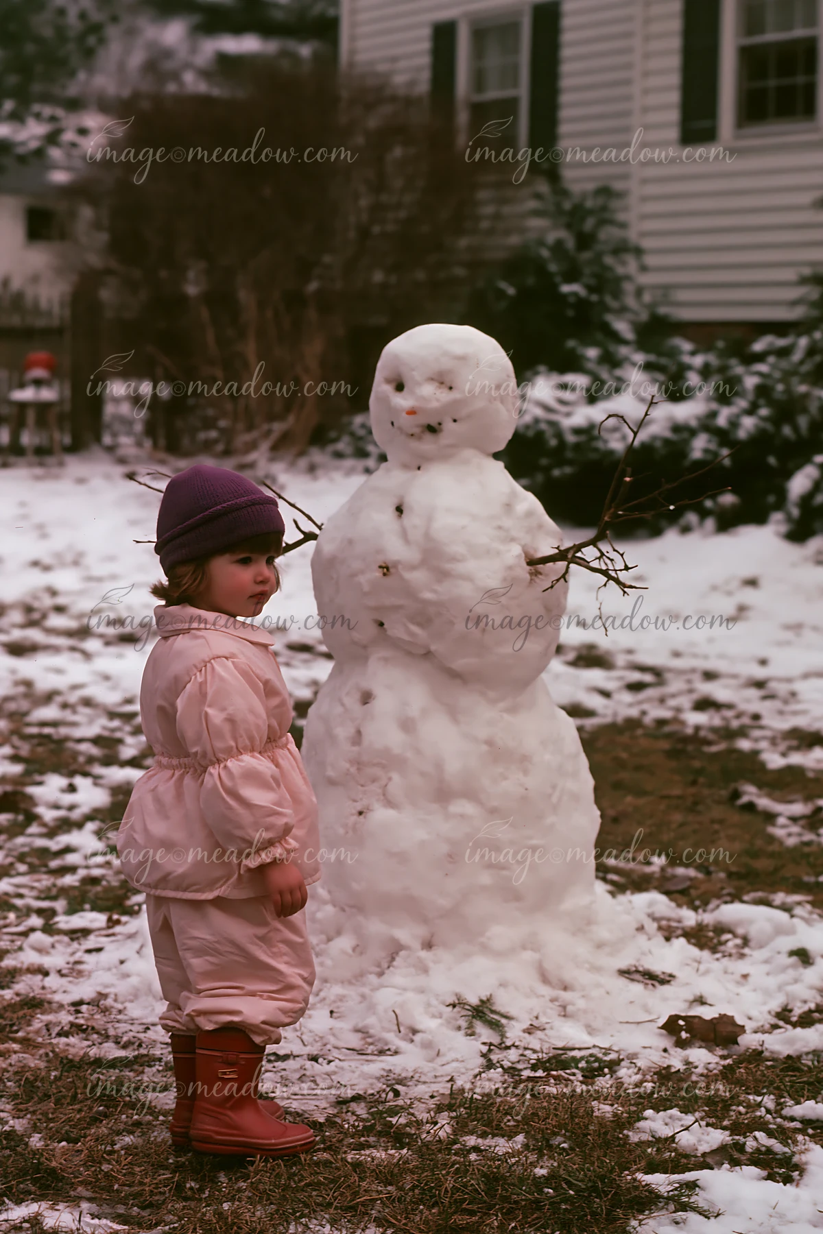 A child stands beside a snowman in a snowy backyard.