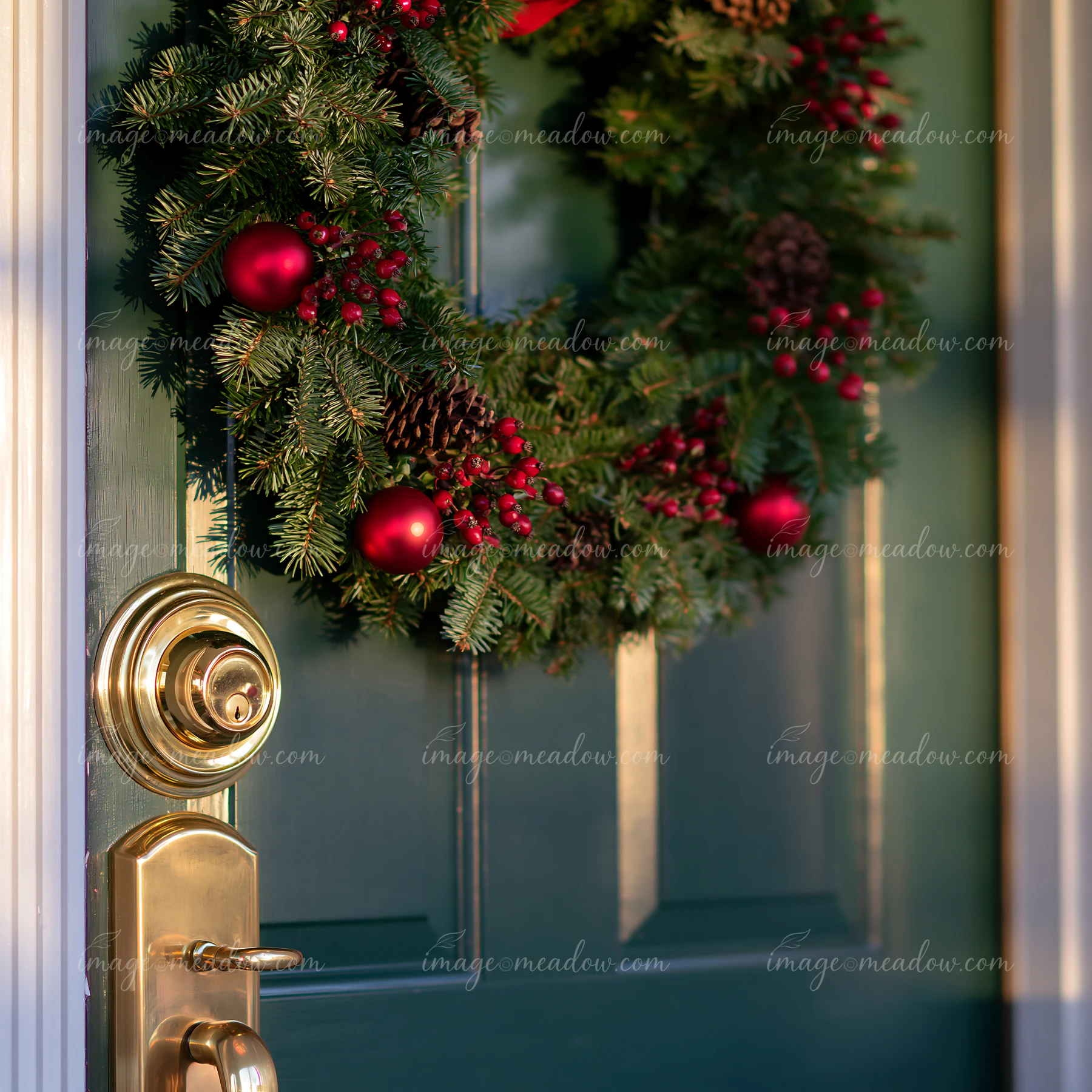A Christmas wreath made of pine and decorated with red ornaments is hung on a green apartment door.