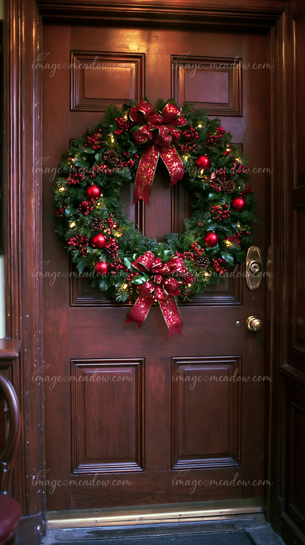 A circular Christmas wreath with red ornaments and ribbons hangs on a wooden apartment door.