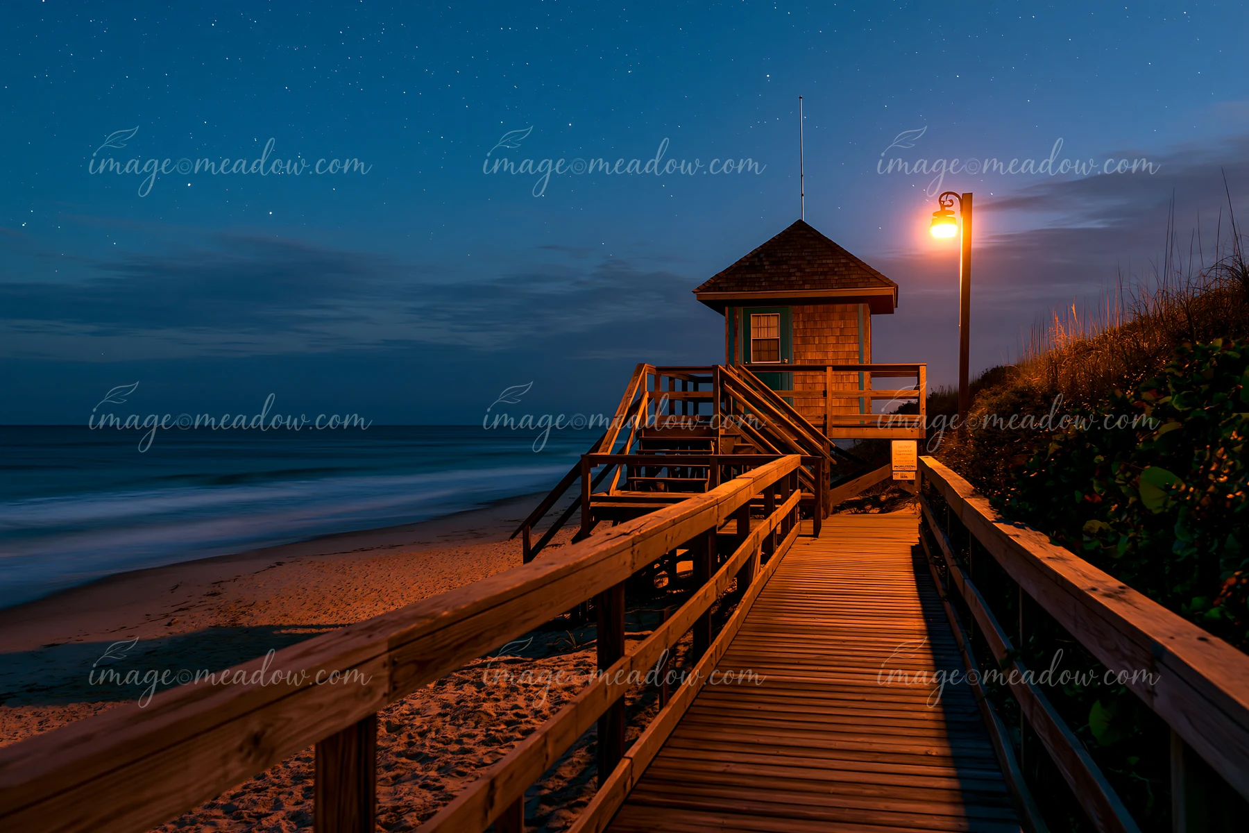 A coastal boardwalk leads to a small lighthouse under a starry night sky.