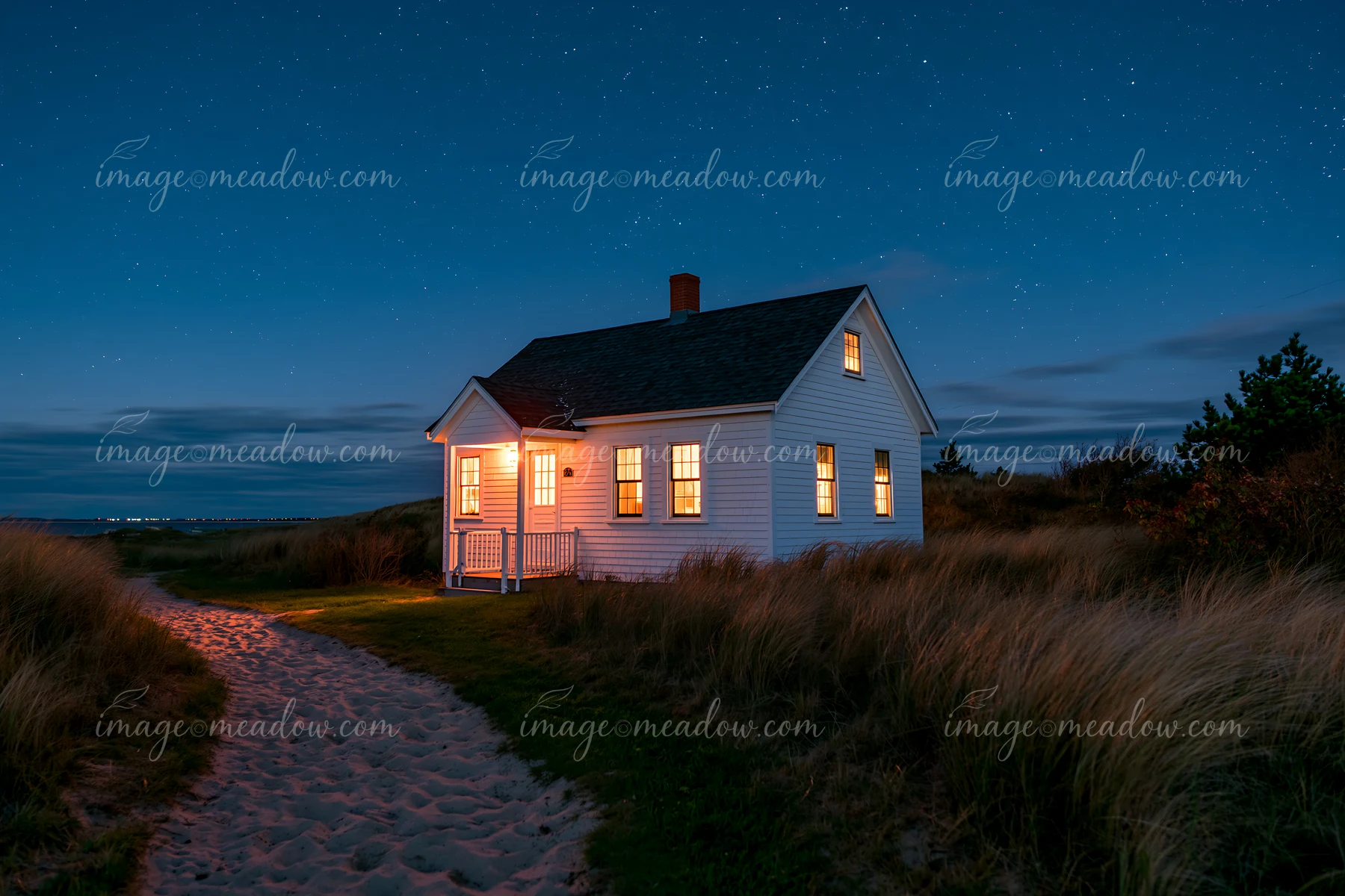 A coastal cottage under stars stands near a sandy path surrounded by grass.
