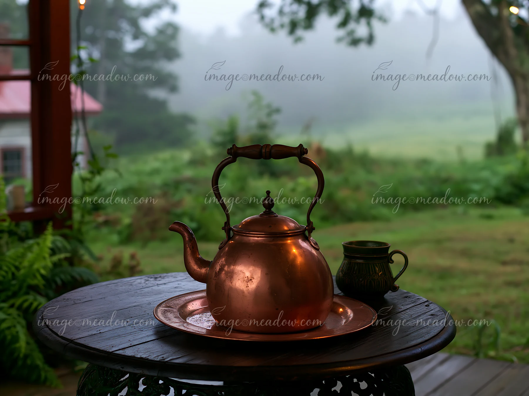 A copper kettle sits on a wooden table near a cottage in the misty countryside.