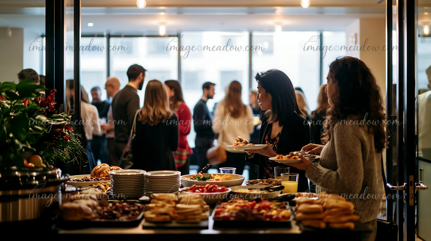 A corporate lunch buffet features diverse dishes as guests fill their plates and socialize in a bright indoor space with large windows.