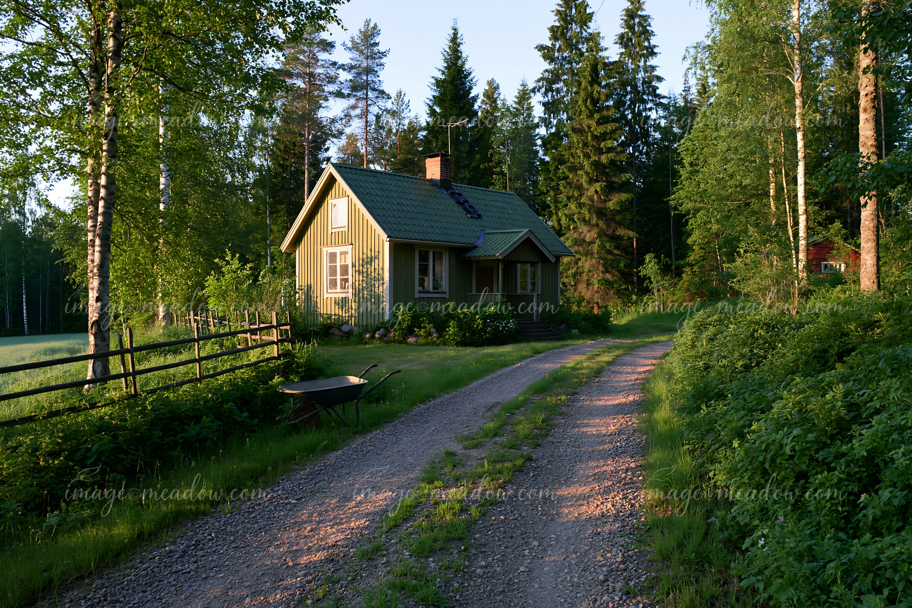 A cottage near forest edge sits beside a gravel path surrounded by trees and shrubs.