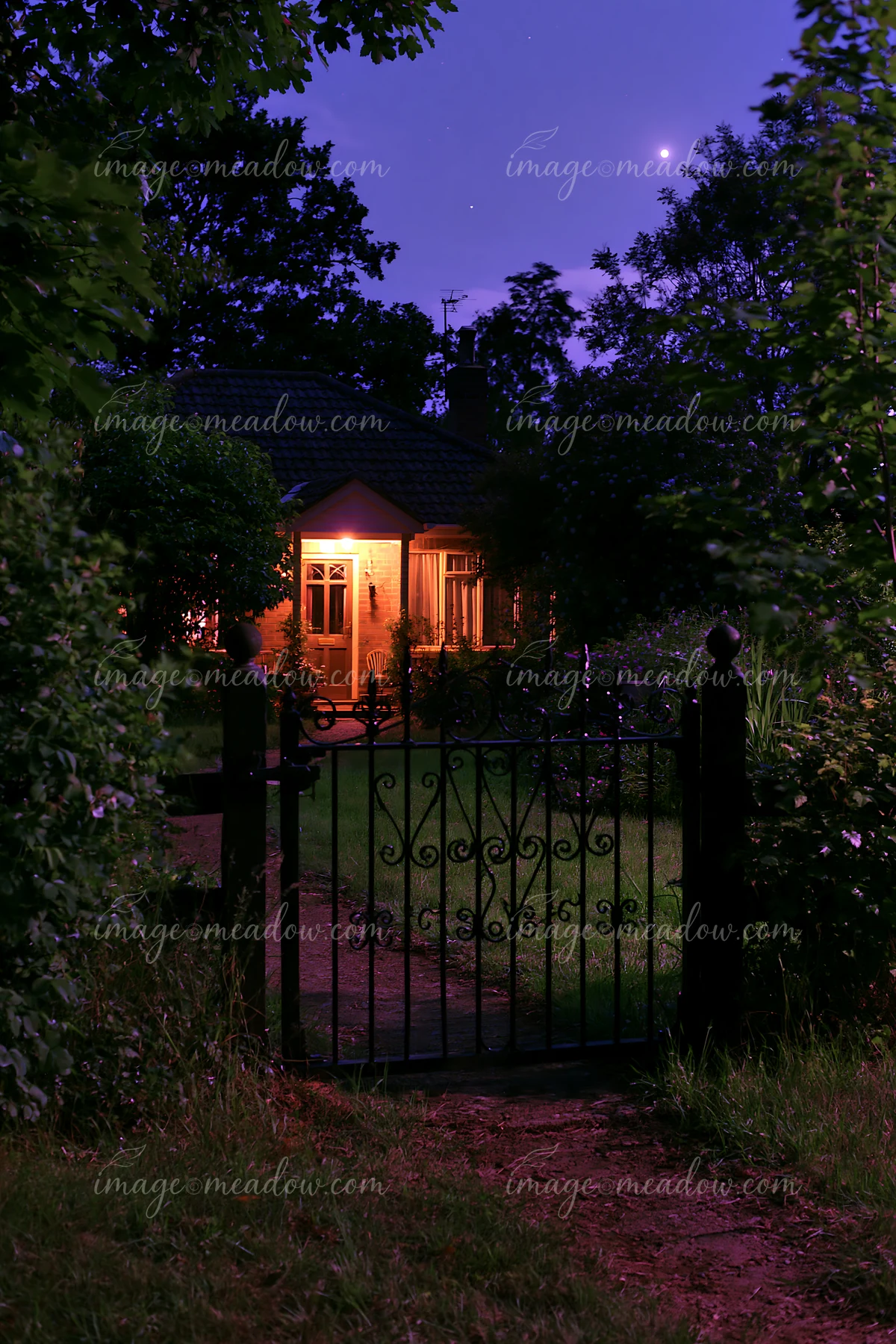 A country home sits behind a decorative gate, softly lit by warm lights as the night sky is clear.