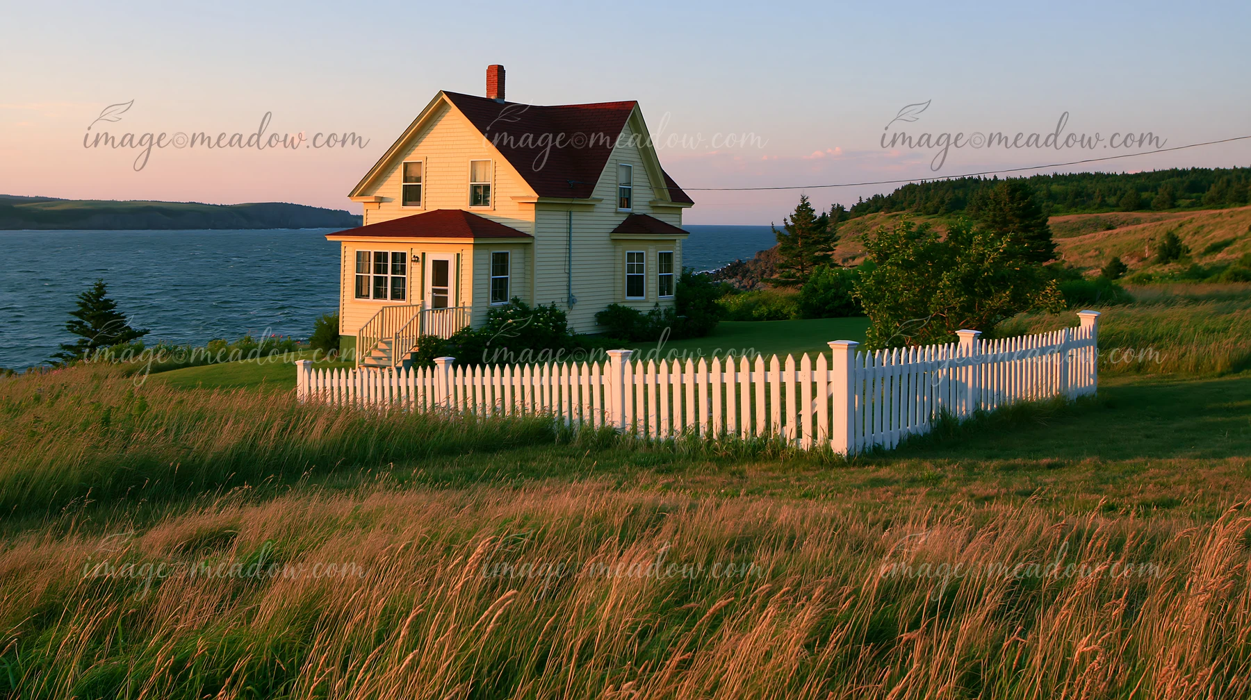A country home with white picket fence sits on a grassy hill beside water.