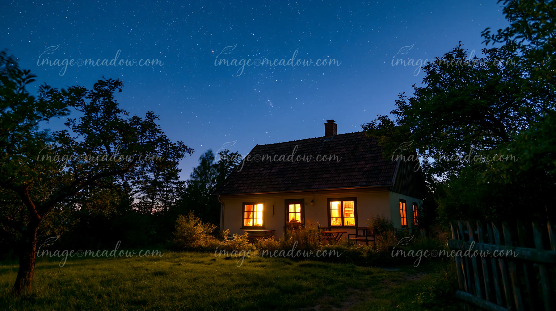 A country house stands under a starry sky, with warm light glowing from its windows, surrounded by trees and a grassy yard.