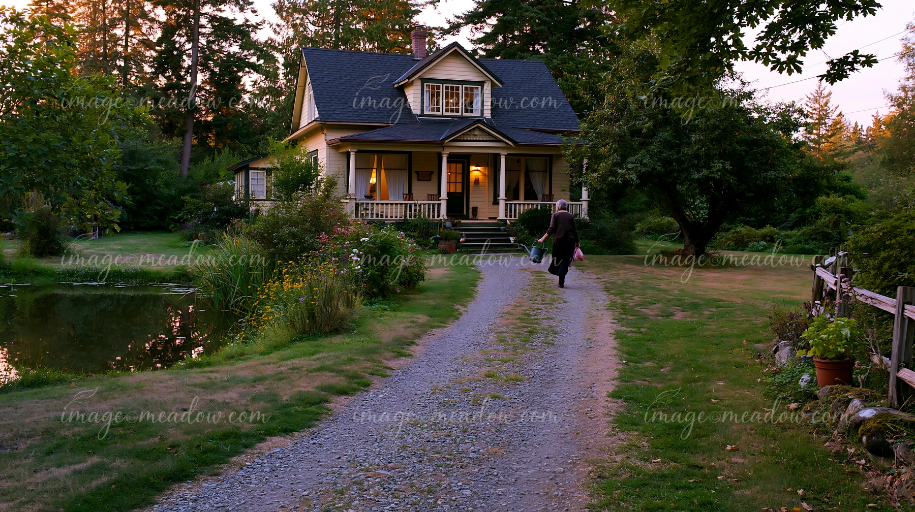 A country house with gravel driveway stands near a pond surrounded by trees.
