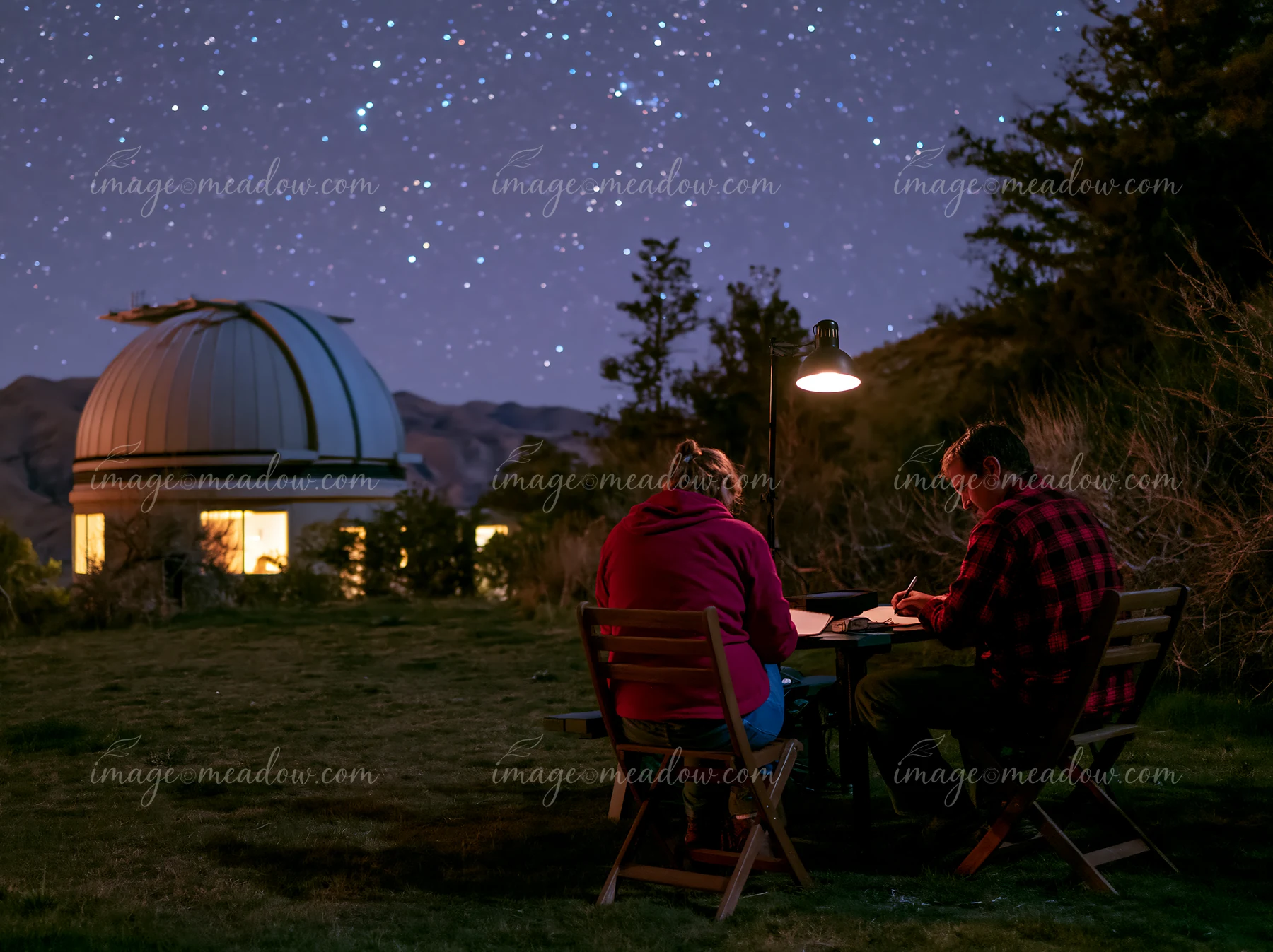 A couple at a remote observatory at night enjoy stargazing while seated at a table outside.