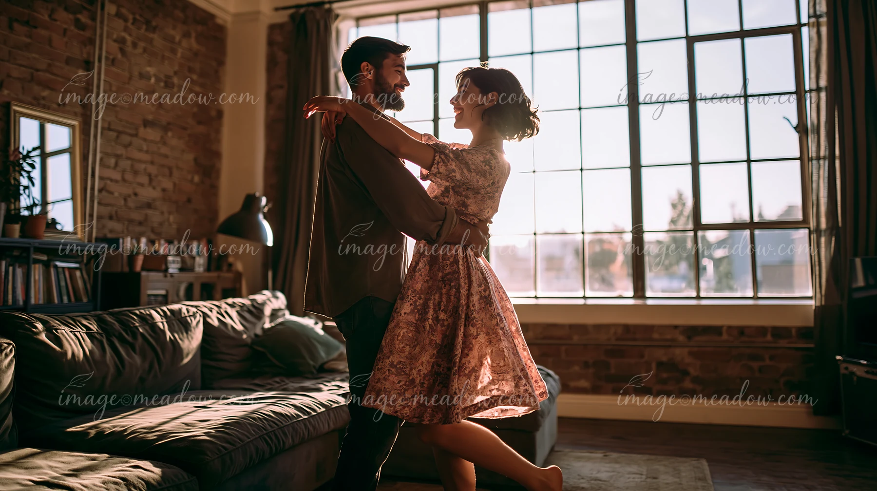 A couple dances barefoot in a living room filled with warm light from large windows.