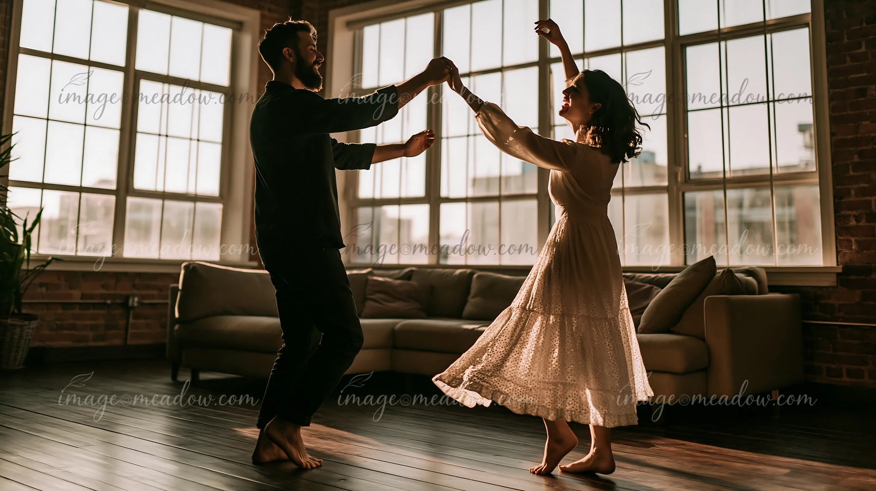 A couple dances barefoot in a living room with large windows, enjoying each other's company.