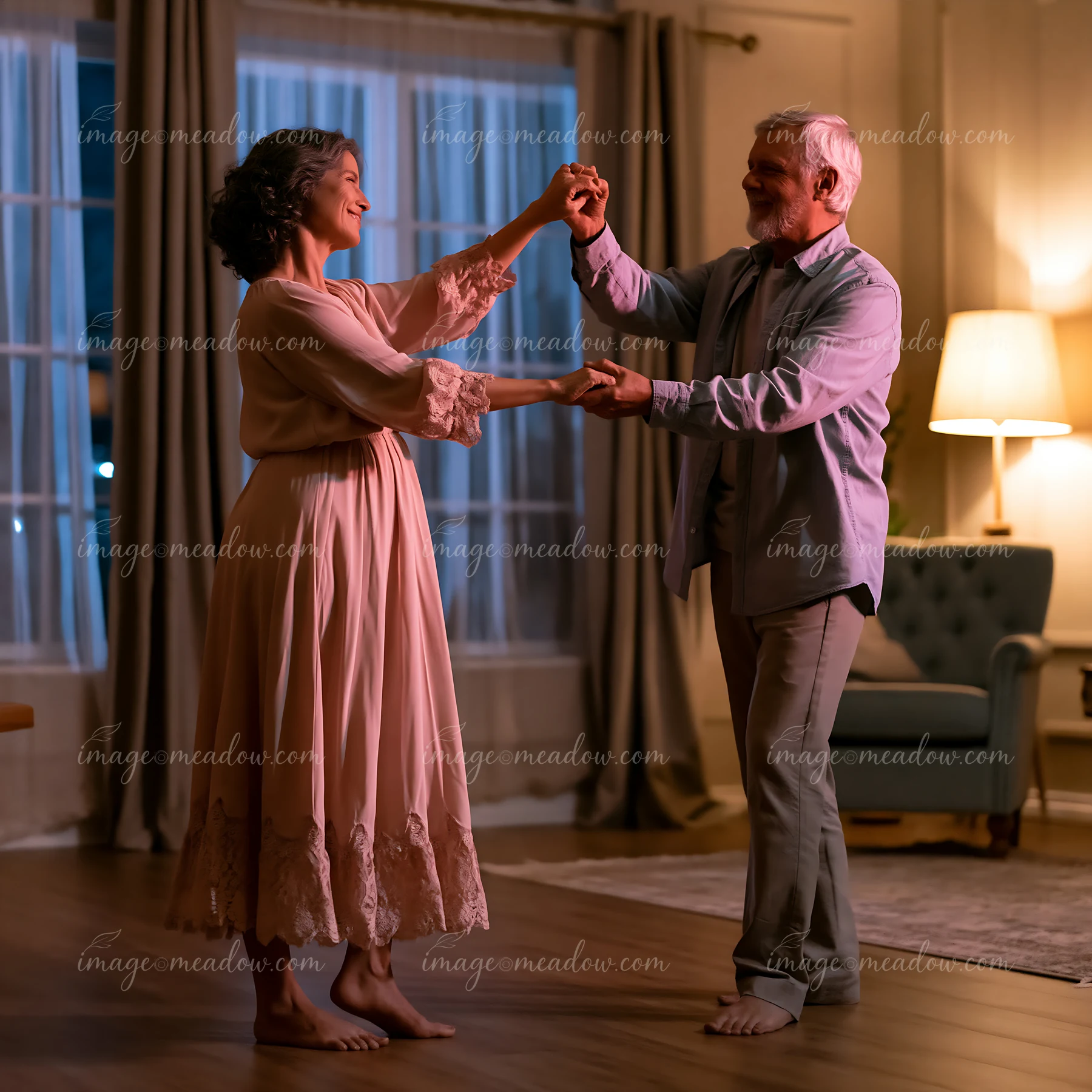 A couple dances barefoot in their living room at night, holding hands and enjoying each other's company.