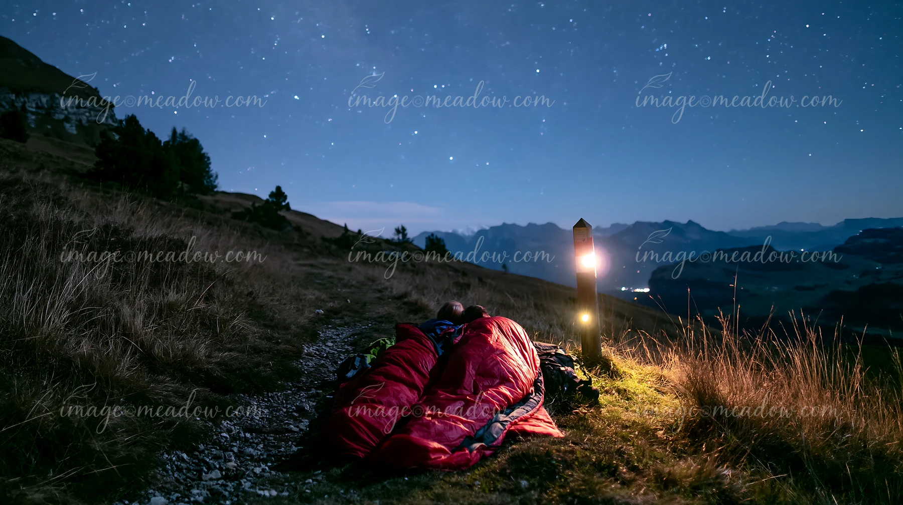 A couple is sharing a sleeping bag beneath a starry sky on a mountain path.