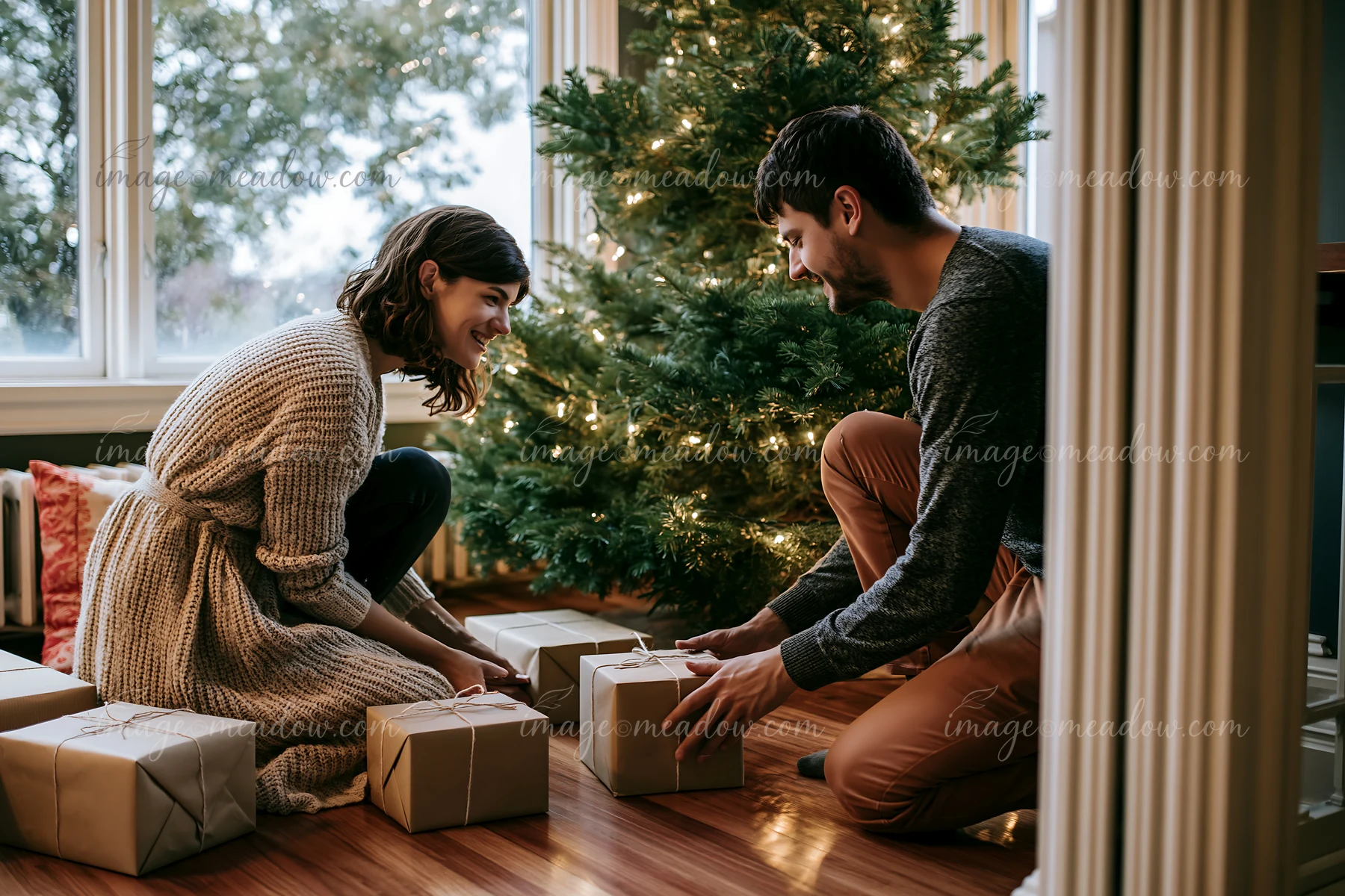 A couple is wrapping gifts and arranging them under a Christmas tree in a warm living room.