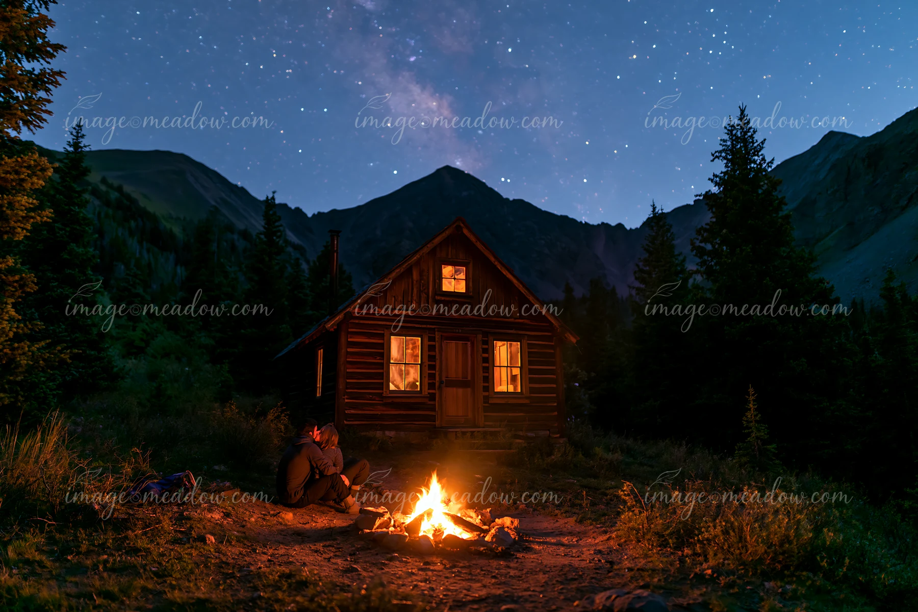 A couple kisses beside a campfire under stars near a wooden cabin in the mountains.