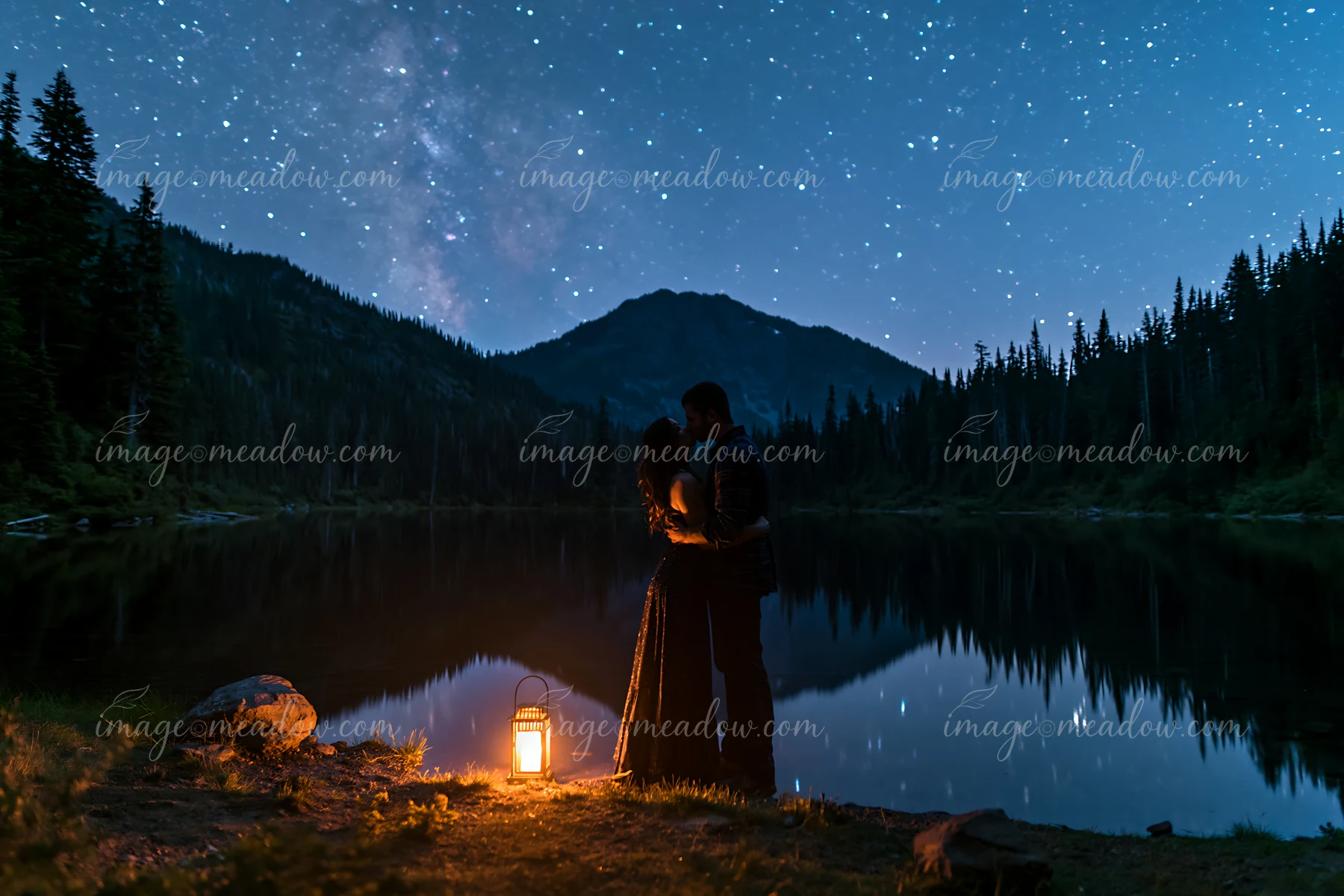 A couple kissing under stars beside a lake surrounded by trees and mountains.