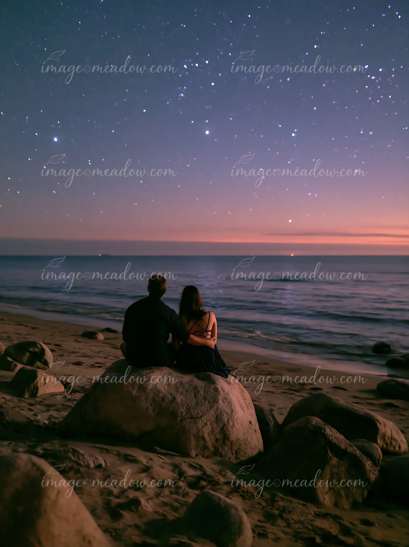 A couple on a beach under stars sits on a large rock, gazing at the starry sky and ocean waves.