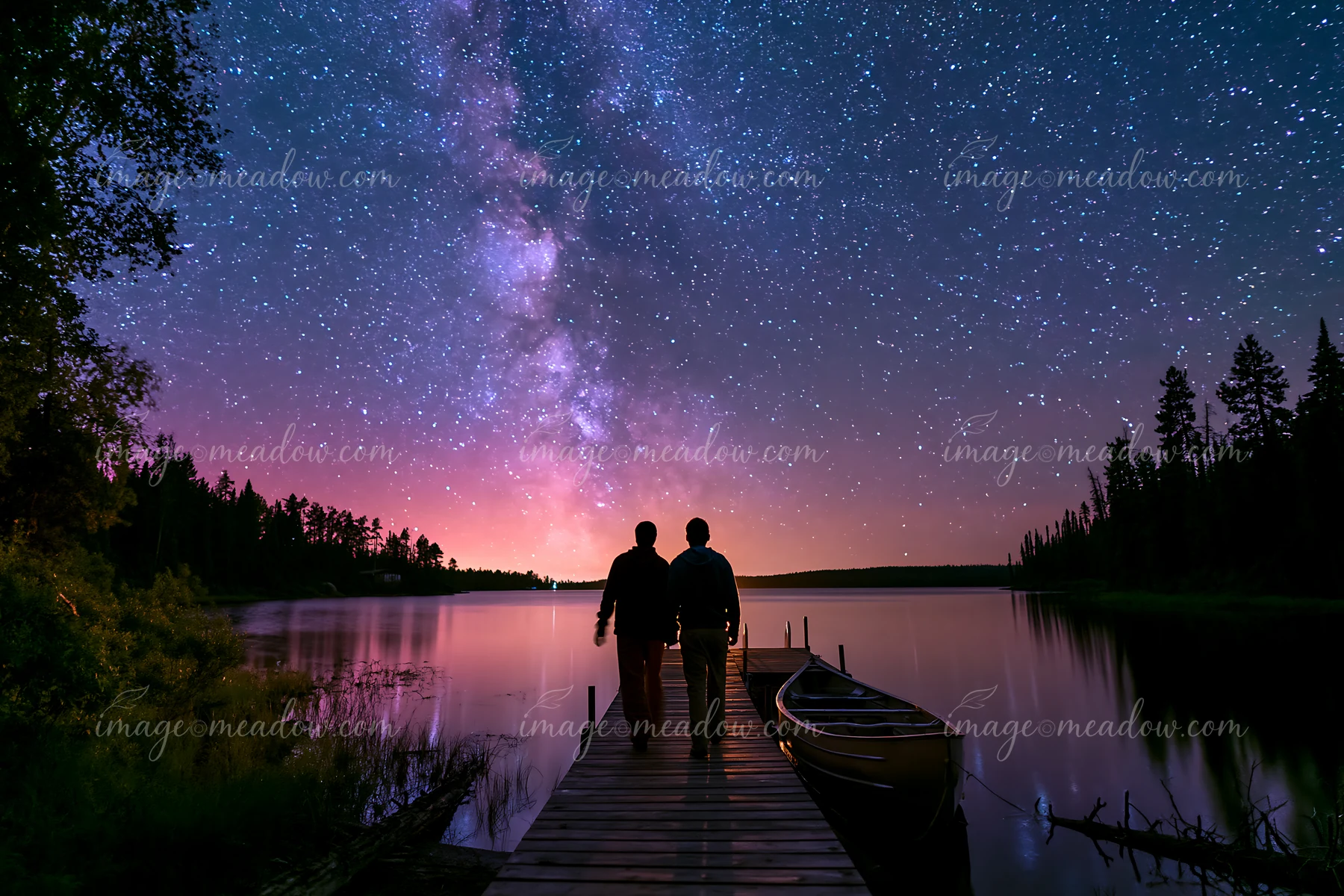 A couple on a dock beneath the Milky Way enjoys a night by the calm water.