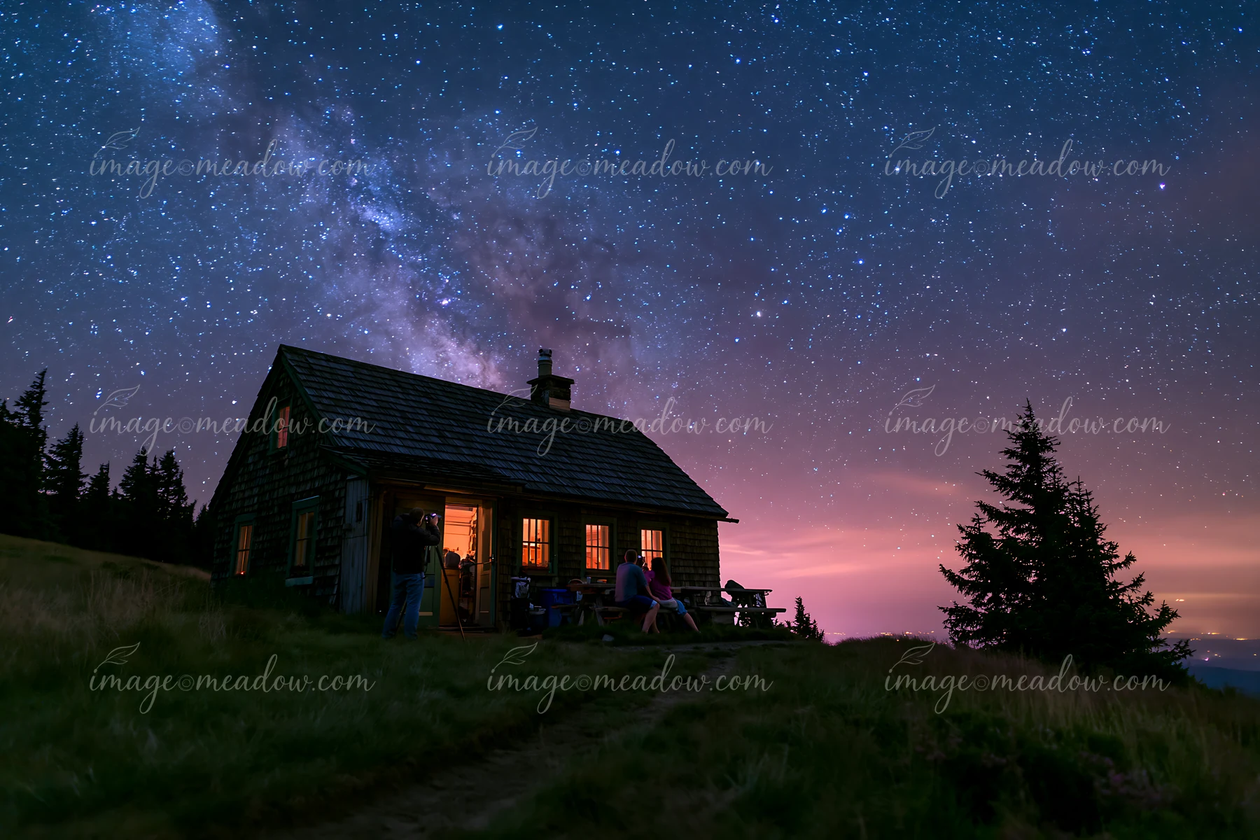 A couple photographing Milky Way at night near a cabin surrounded by trees.