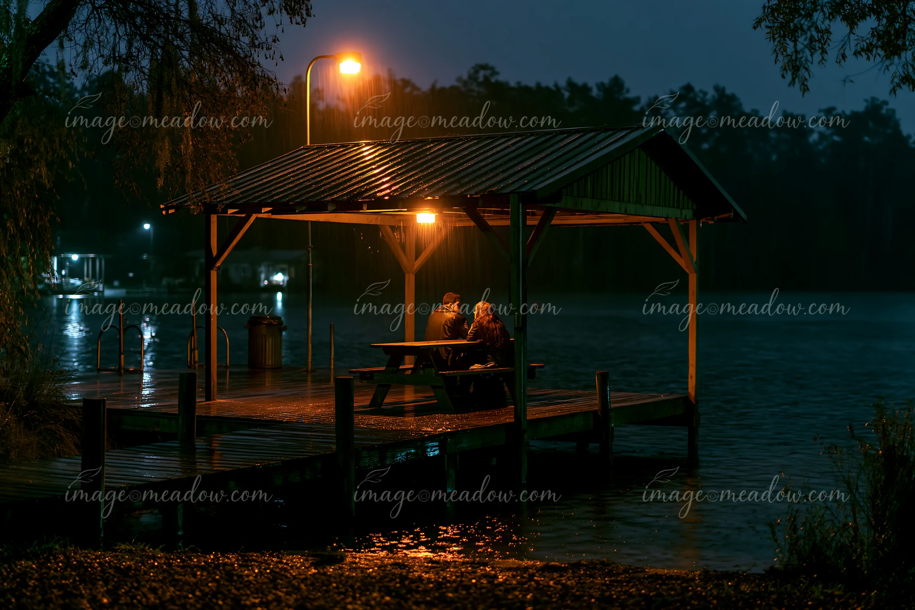 A couple relaxing beside a lake at night sits on a dock.