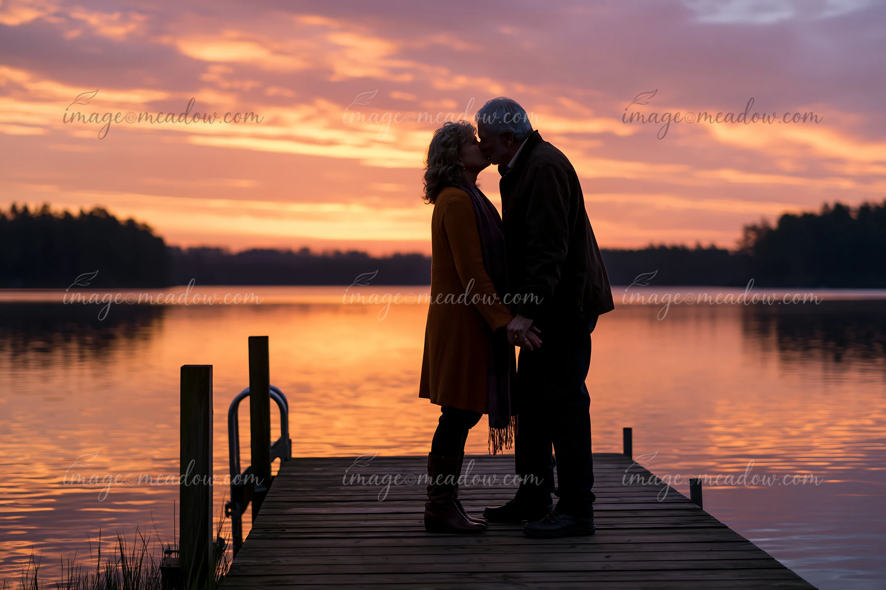 A couple shares a romantic kiss at sunset on a dock by the lake, holding hands as the sun sets, casting a colorful glow on the water.