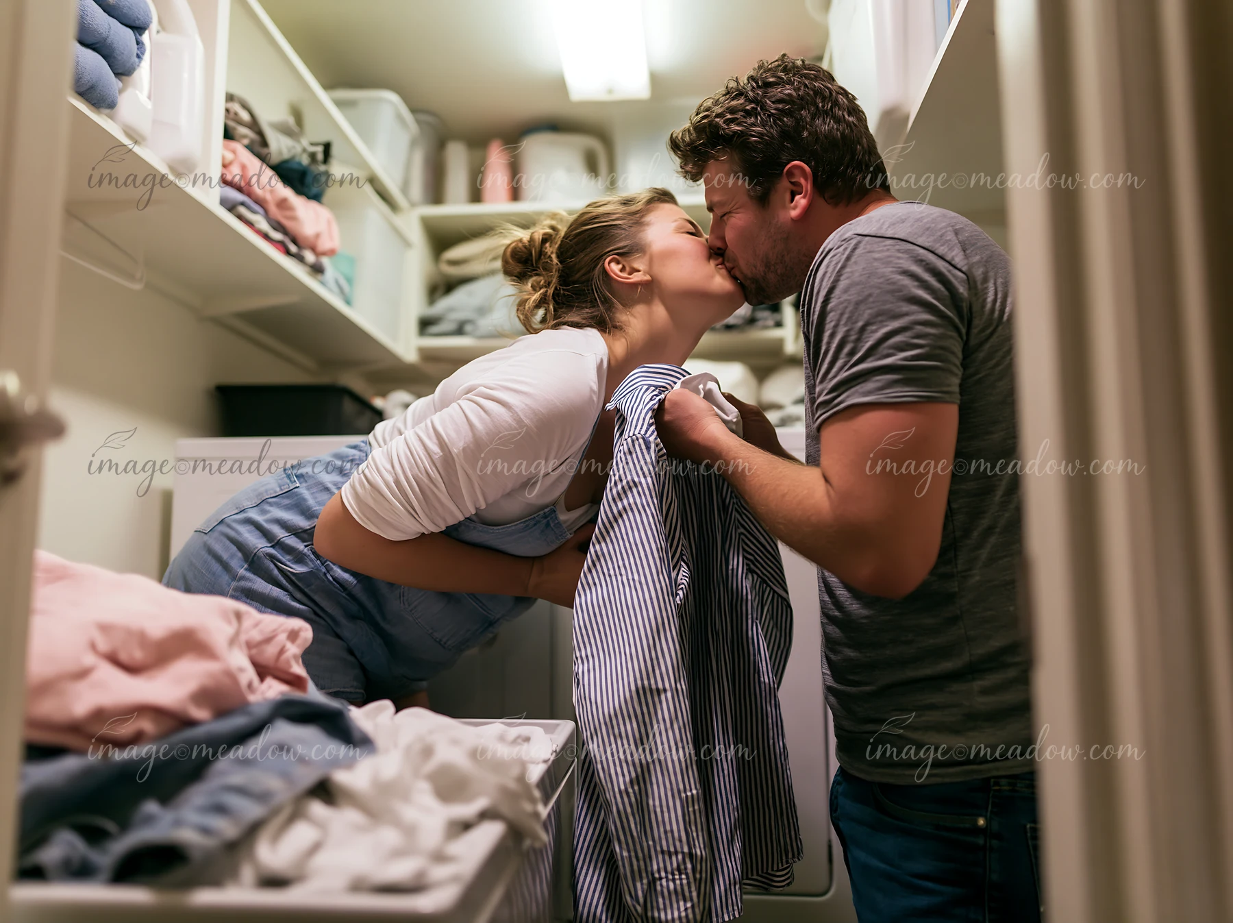 A couple shares a tender kiss while standing in a laundry room filled with clothes.