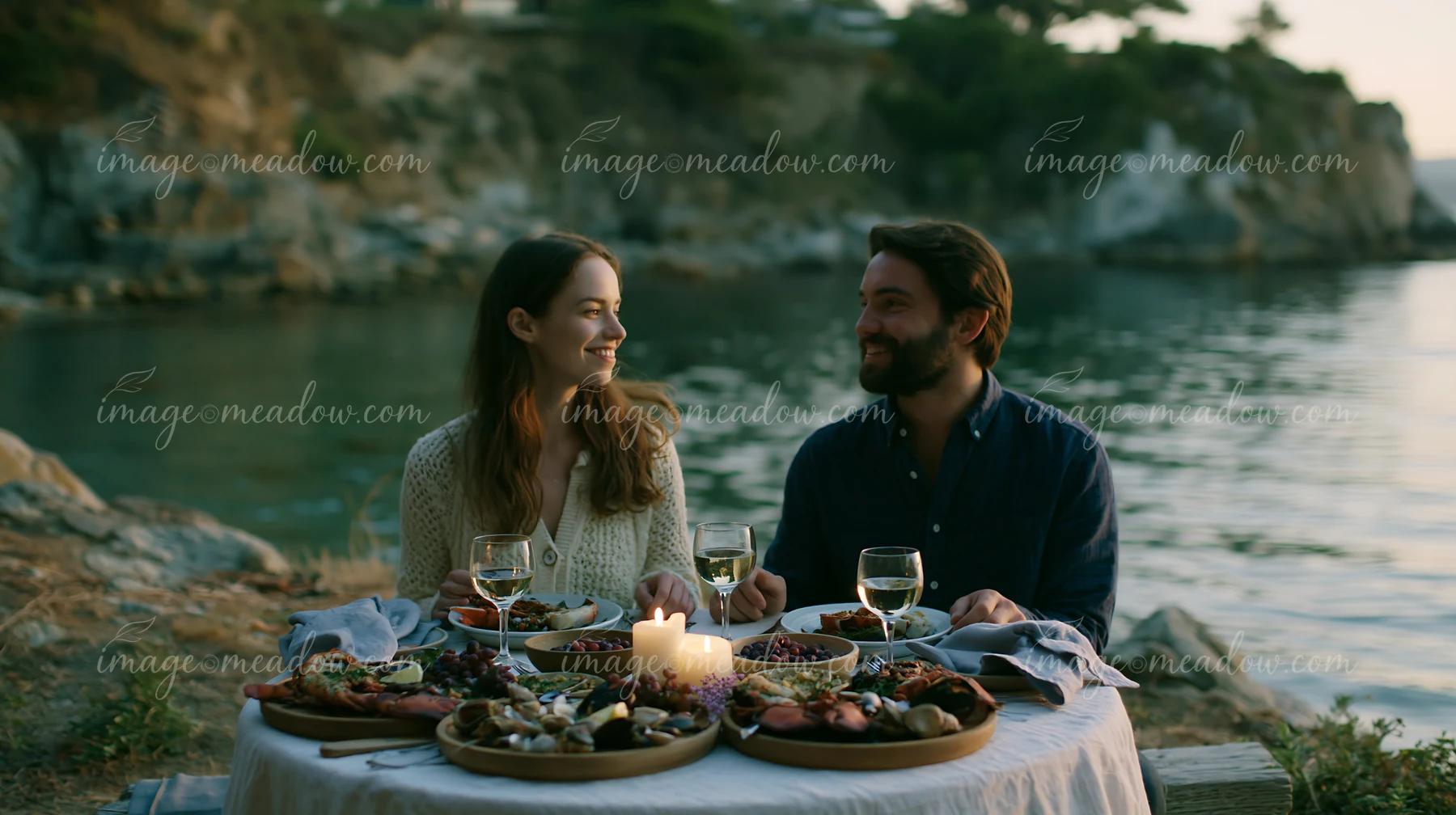 A couple sits at a table on the beach, sharing a buffet meal as the sun sets over the water.