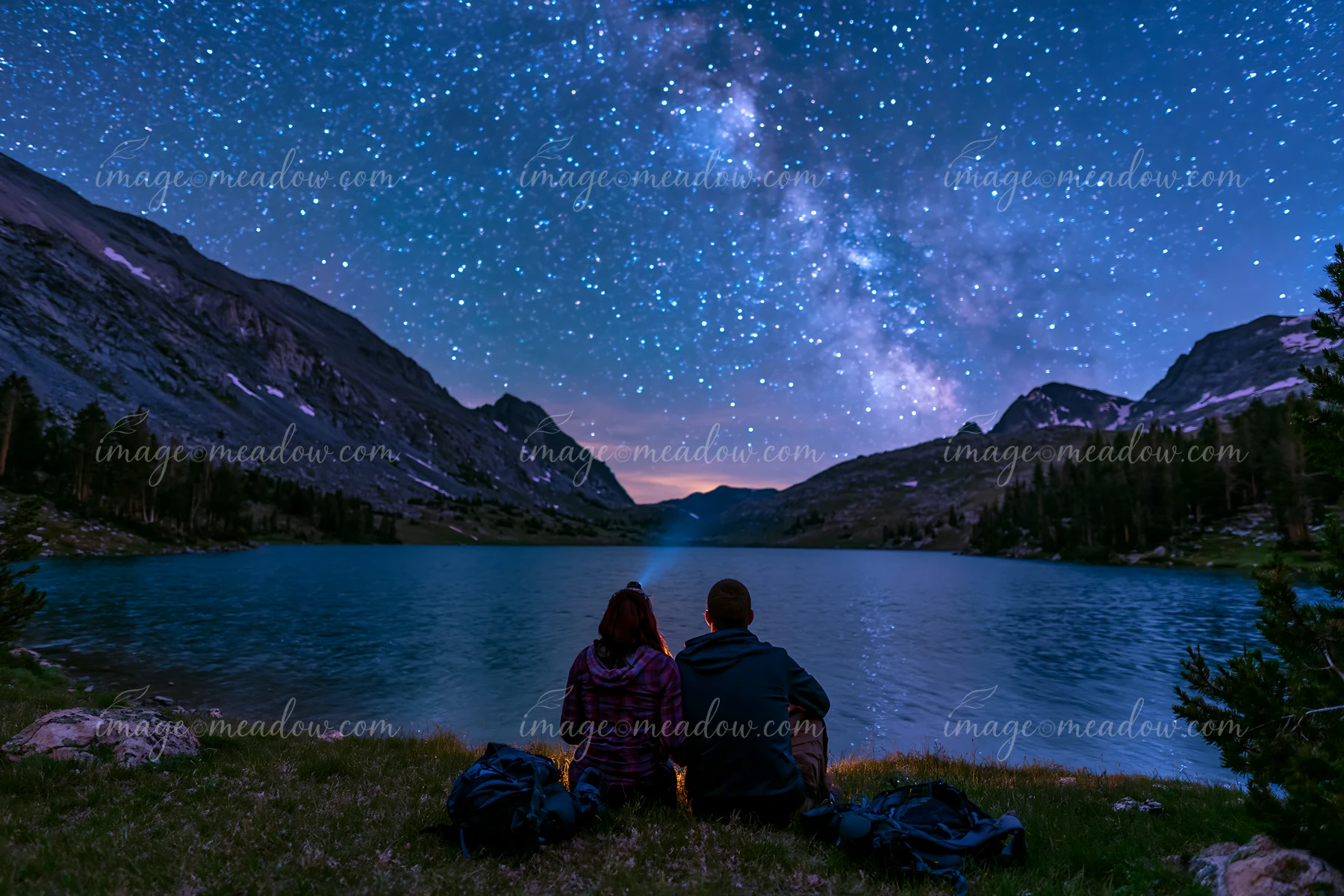 A couple sits by a lake while looking up at a starry night sky filled with stars.