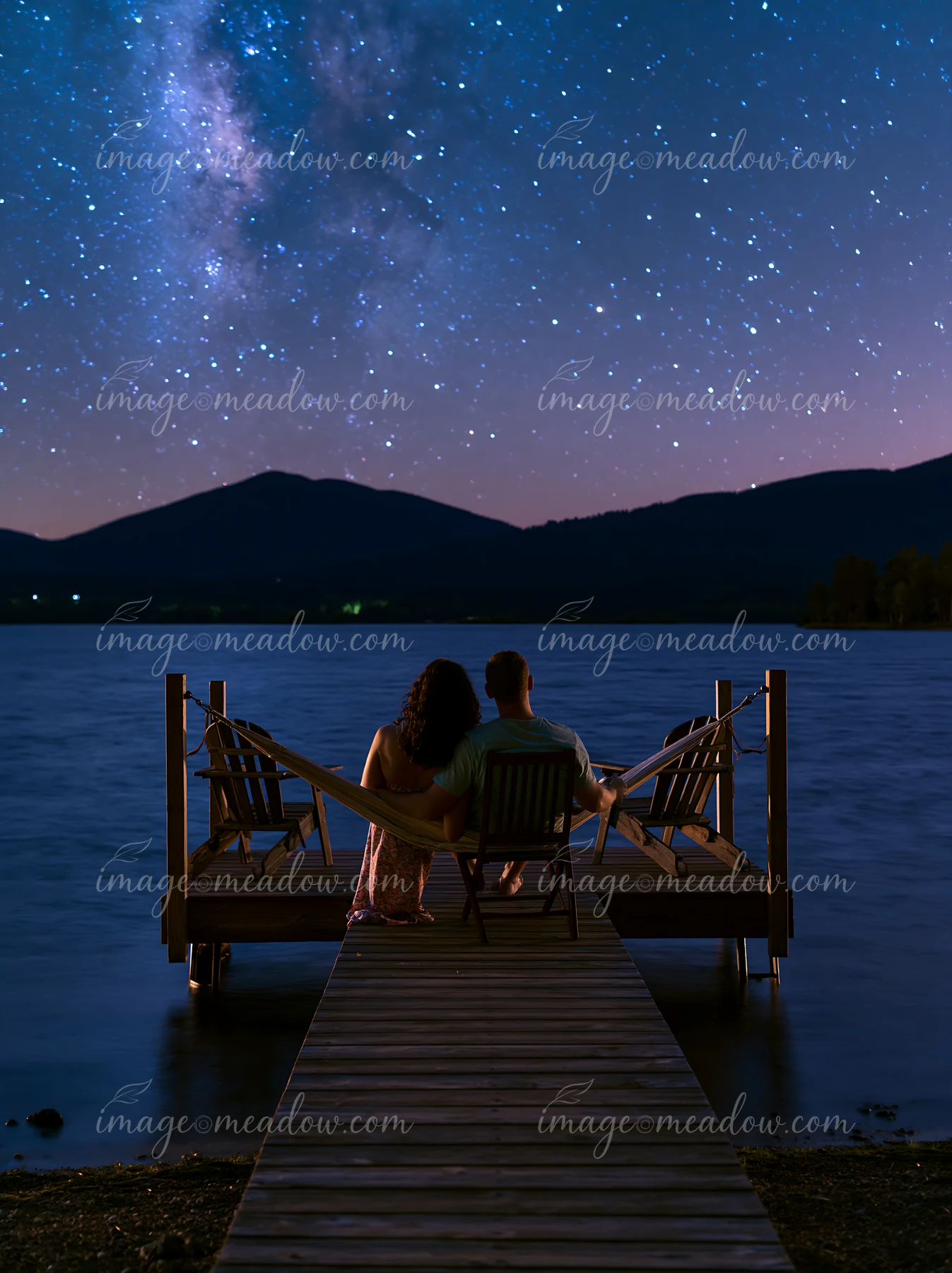 A couple sits in a hammock on a dock by the lake, surrounded by mountains and a clear night sky filled with stars.