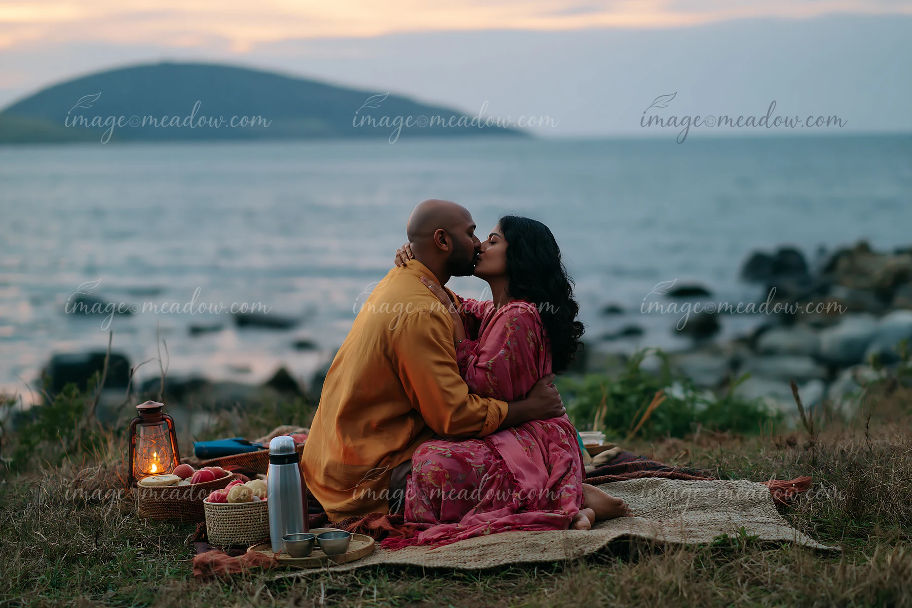 A couple sits on a blanket, sharing a romantic kiss by the water during sunset.