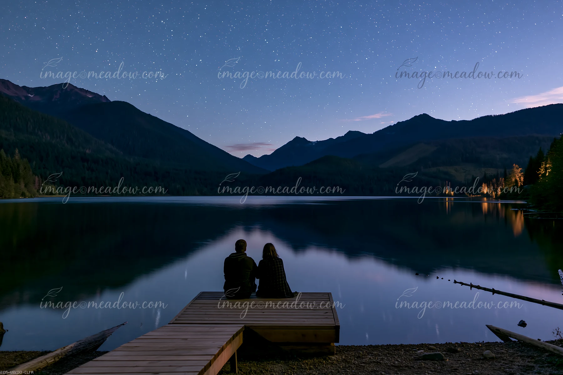 A couple sits on a dock under stars near a mountain lake, enjoying the night sky reflected in the calm water.