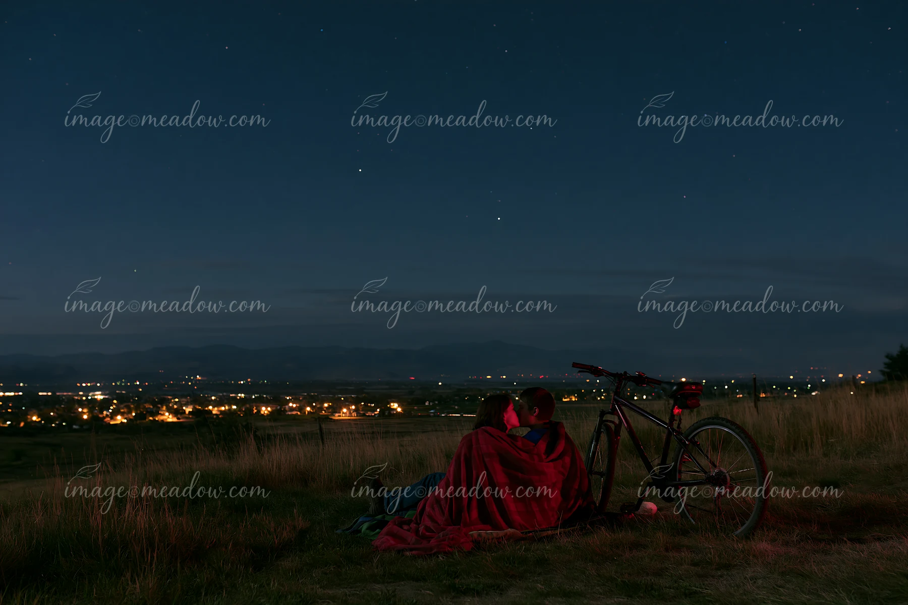 A couple sits on the grass at night, wrapped in a blanket, sharing a kiss under the stars.