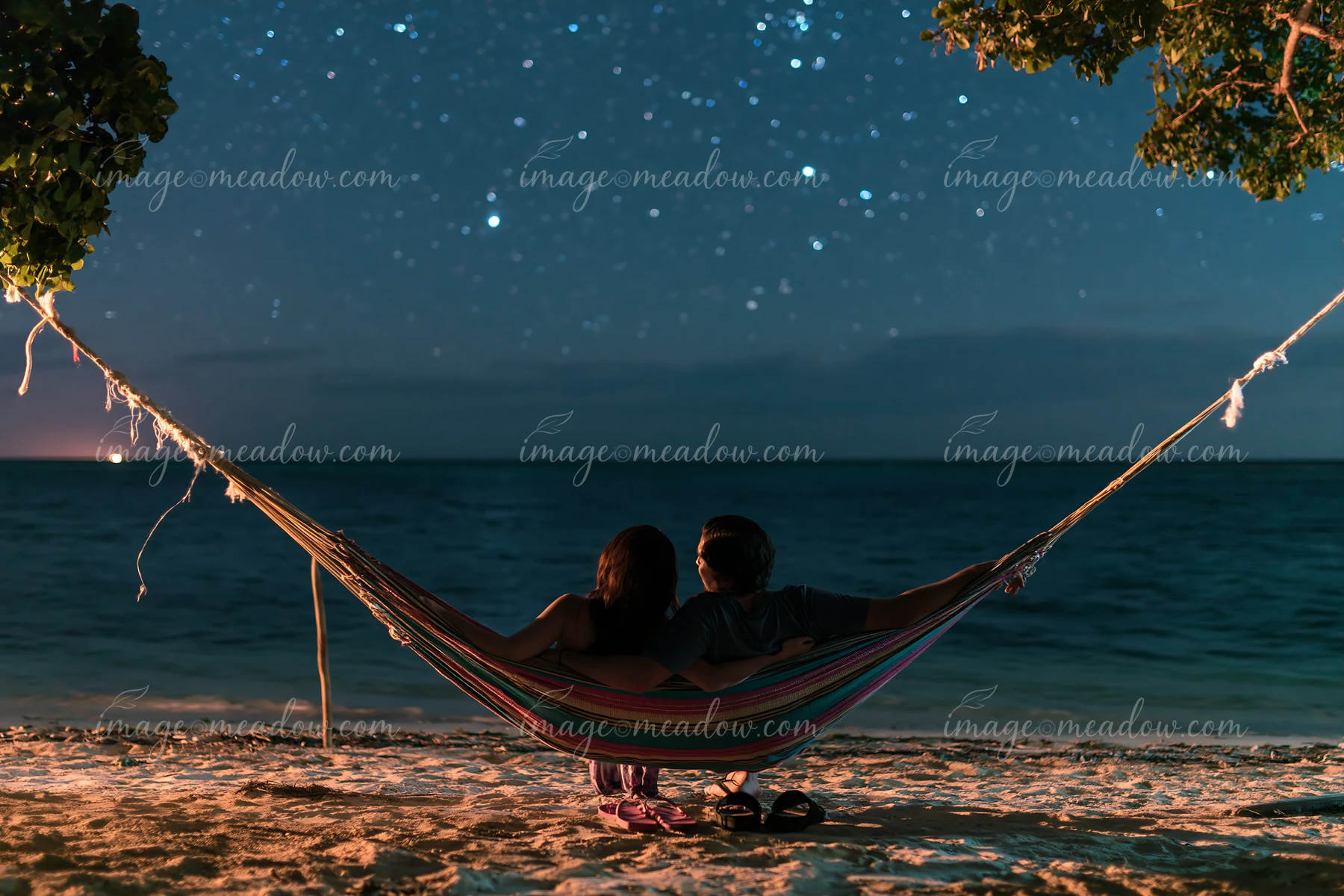 A couple sitting in a hammock under stars relaxes together on the beach, with the ocean in the background.