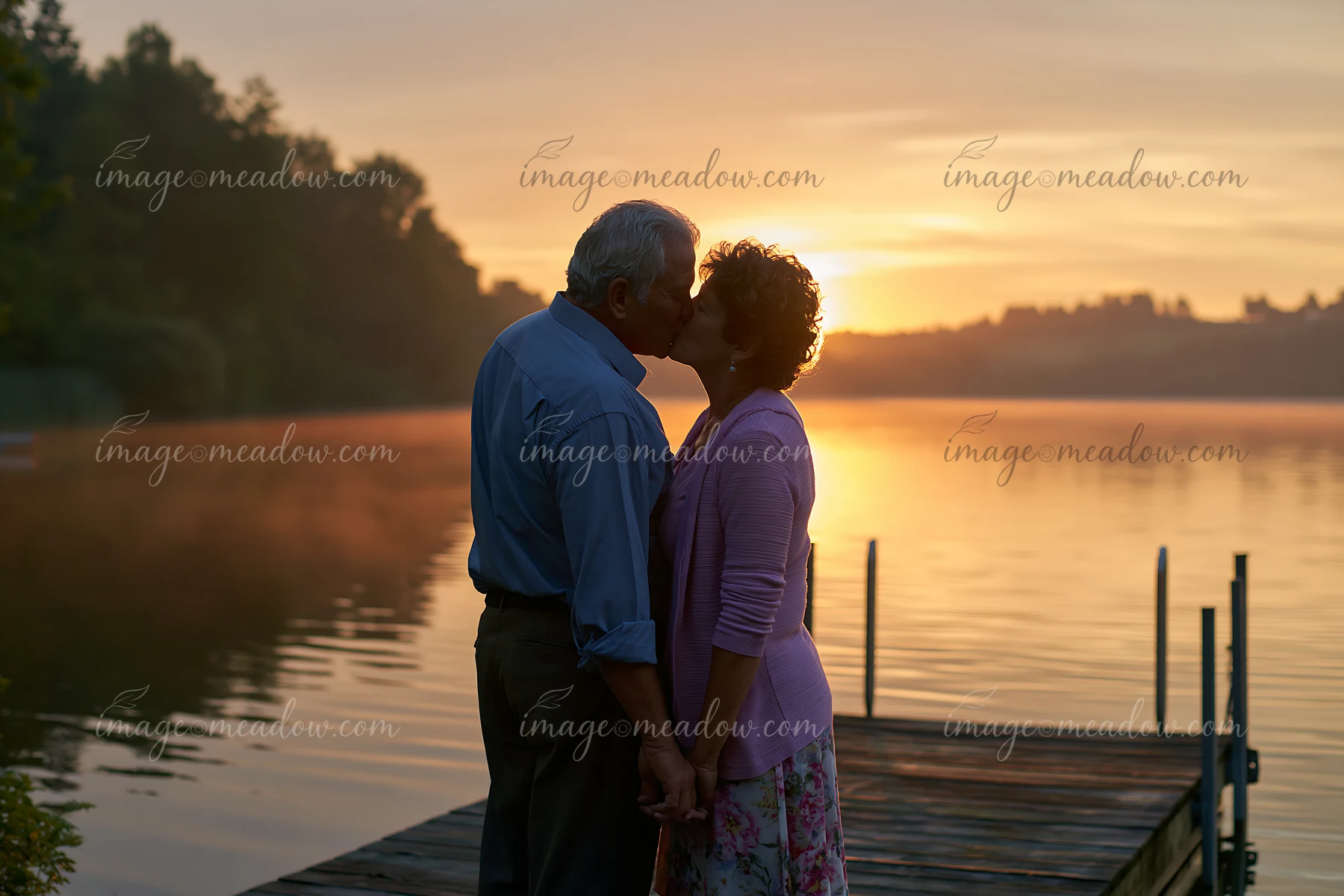 A couple stands on a pier at the lake, sharing a kiss as the sun sets in the background.