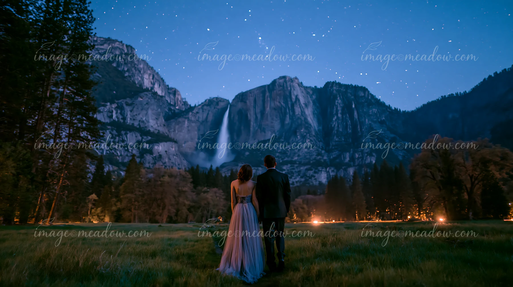 A couple stands together, looking at a waterfall under a starry sky in a forest clearing.