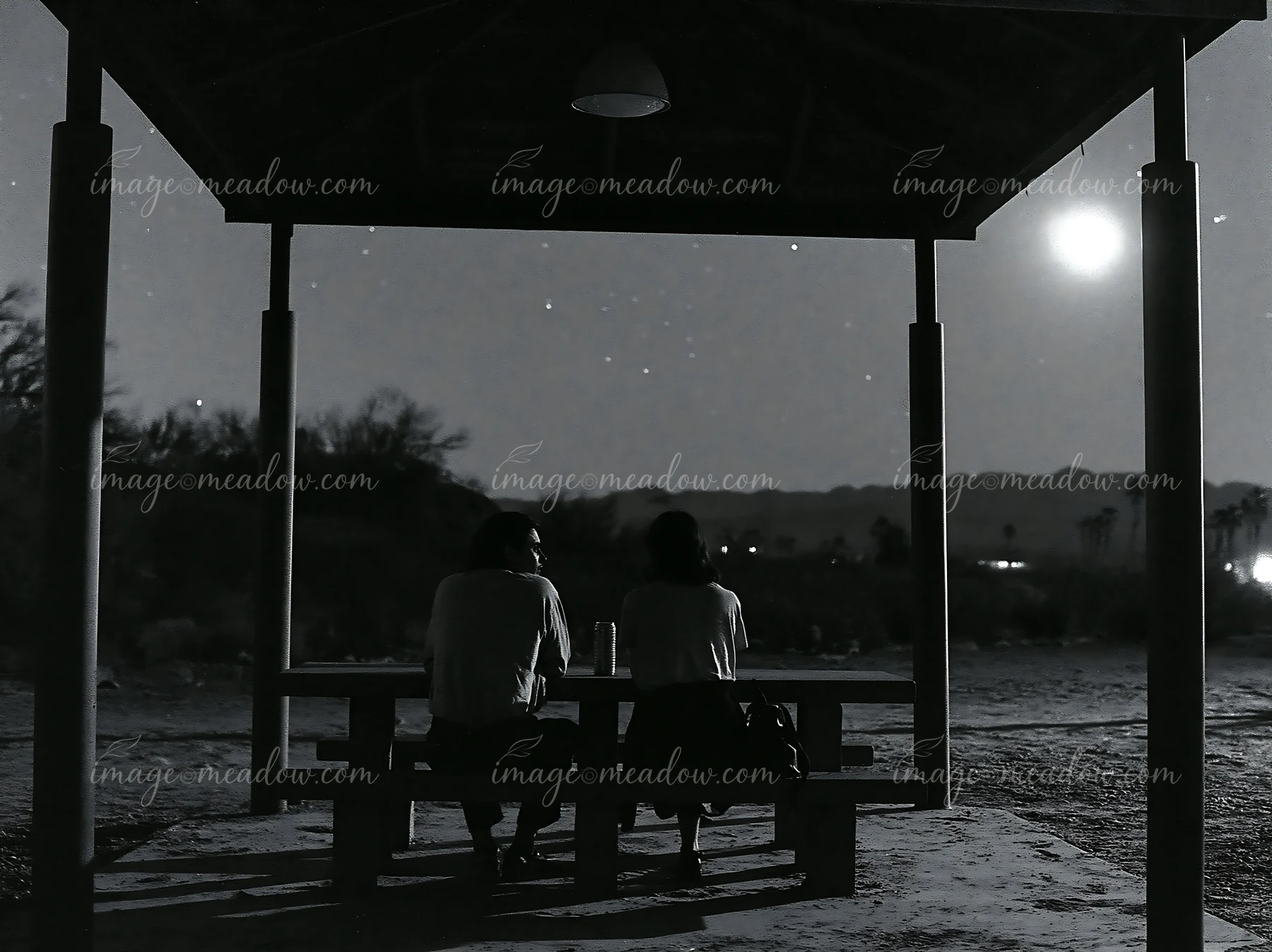 A couple stargazing in a desert at night sits on a bench beneath a shelter.