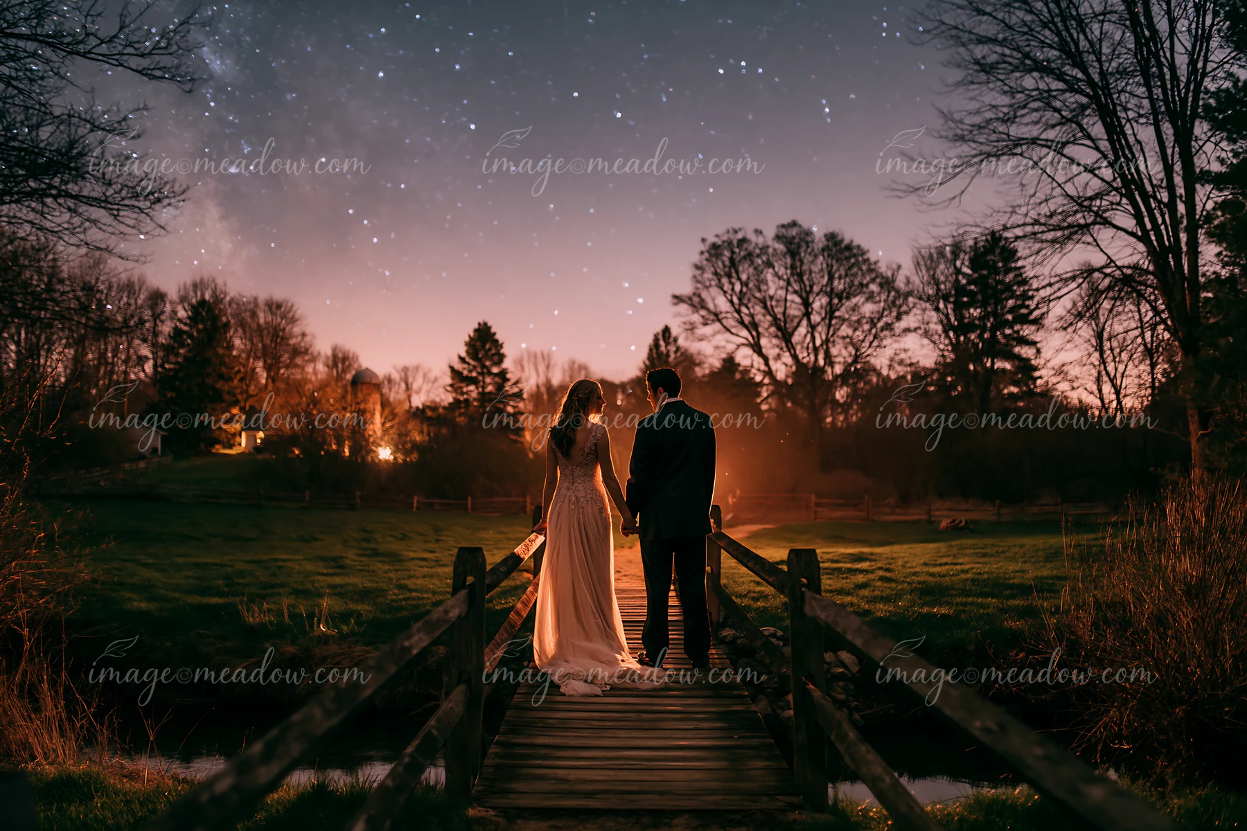 A couple under stars in a rural backyard walks hand in hand on a wooden bridge surrounded by trees and grass.