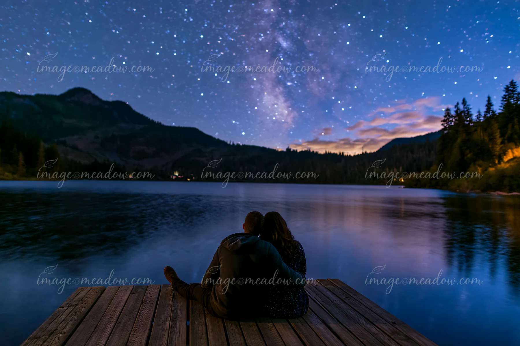 A couple under stars near a mountain lake relaxes on a wooden dock.