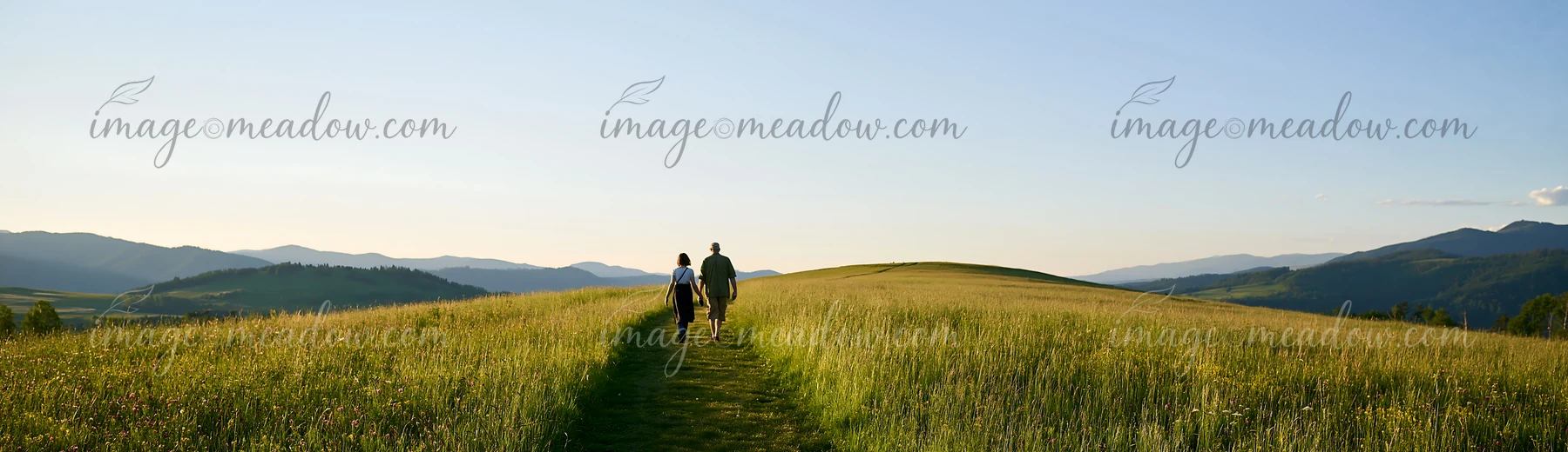 A couple walks along a grassy path in a meadow, surrounded by hills and mountains, enjoying the clear sky.