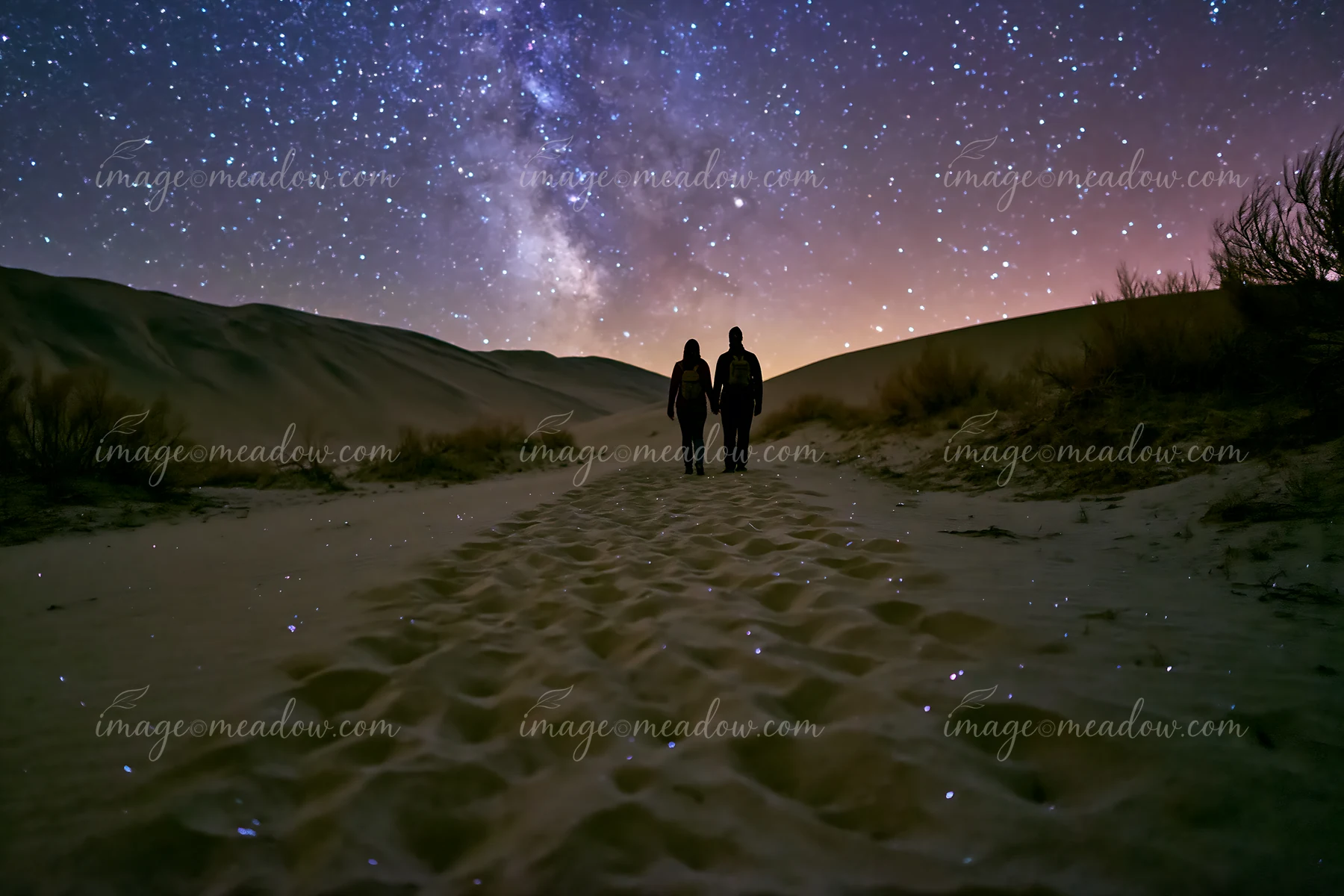 A couple walks on a sandy path in a desert under a bright starry sky.