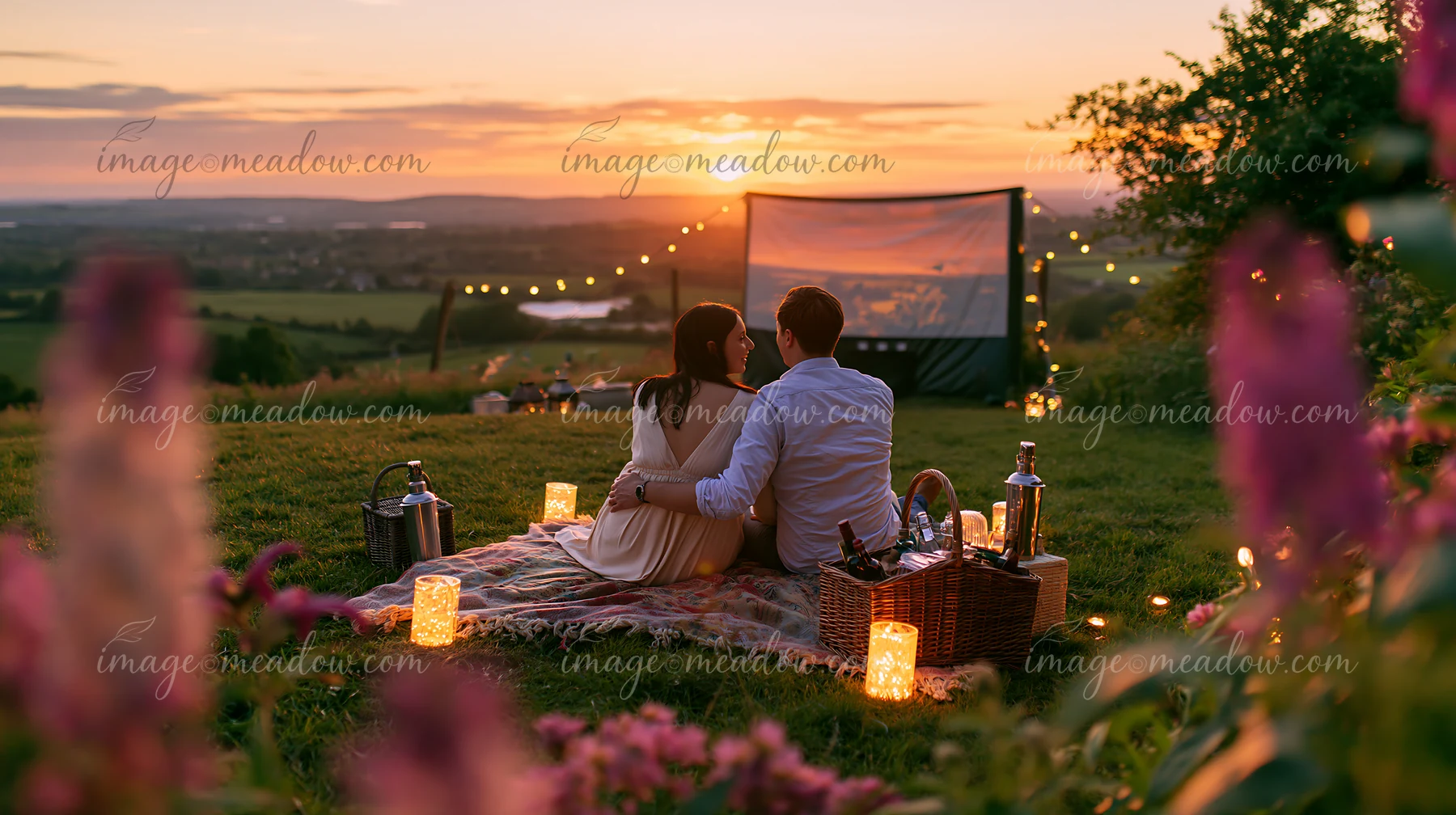 A couple watches an outdoor movie together on a blanket at sunset, surrounded by lanterns, string lights, and picnic baskets.