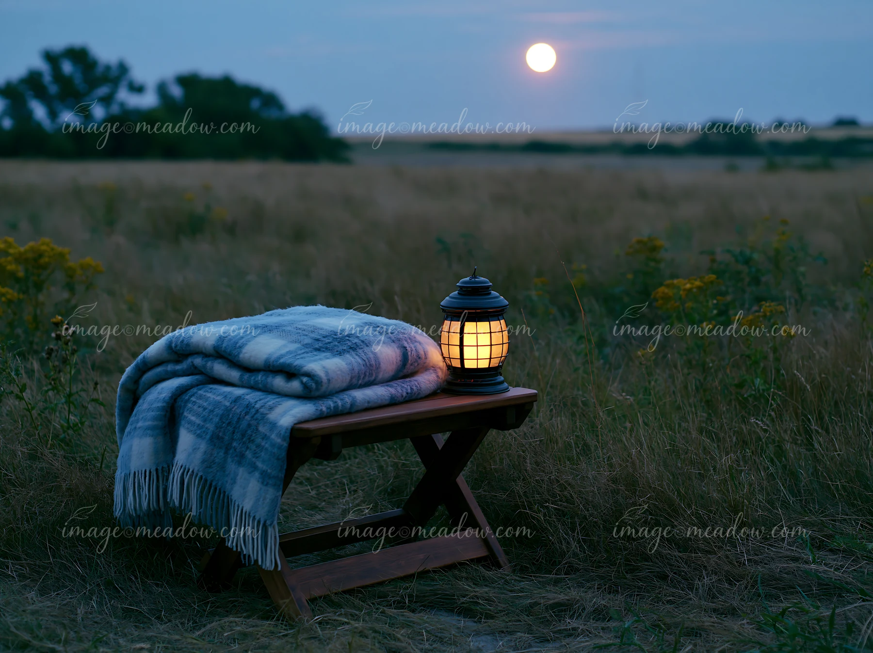 A cozy blanket rests on a wooden stool in a meadow beneath a full moon.