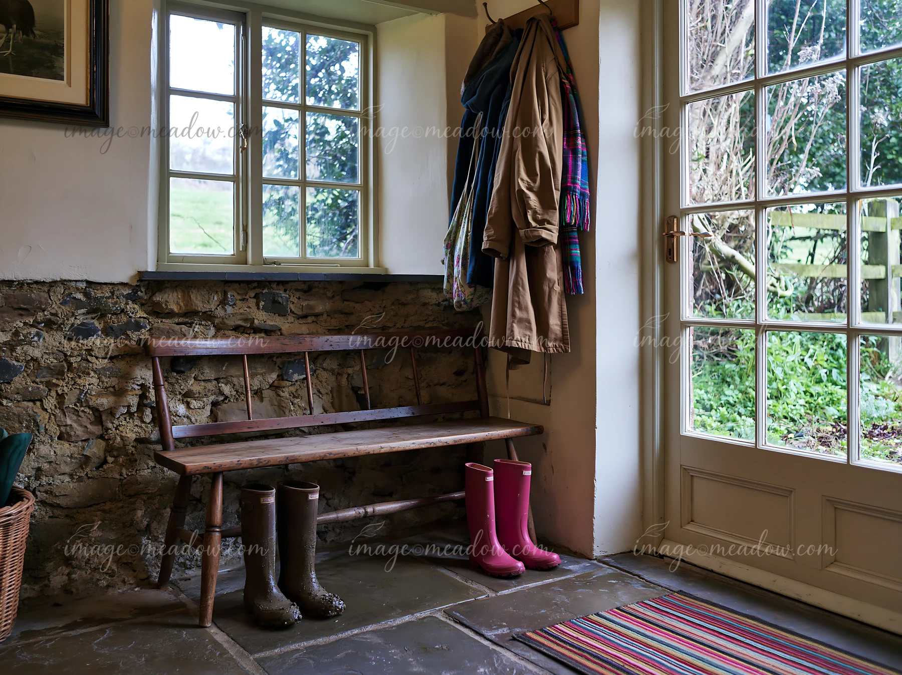 A cozy farmhouse interior with an exposed stone wall showcases a wooden bench and colorful rain boots.