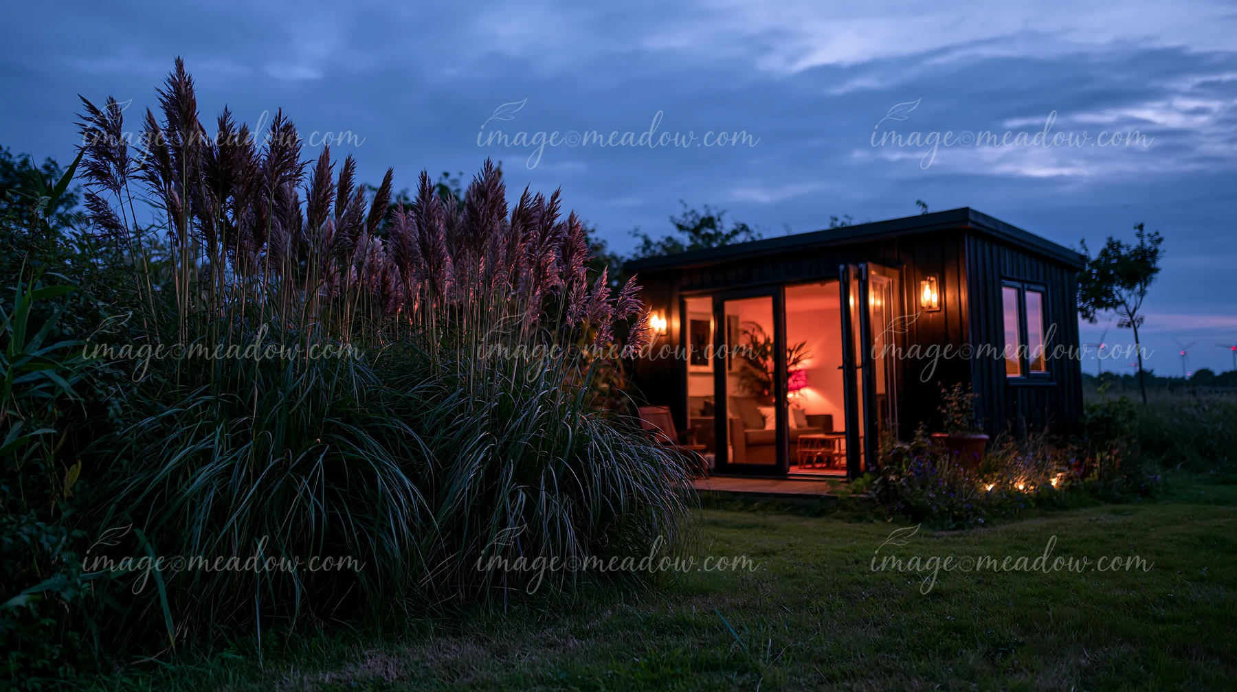 A cozy garden house displays warm lights within, surrounded by tall grass under an evening sky.