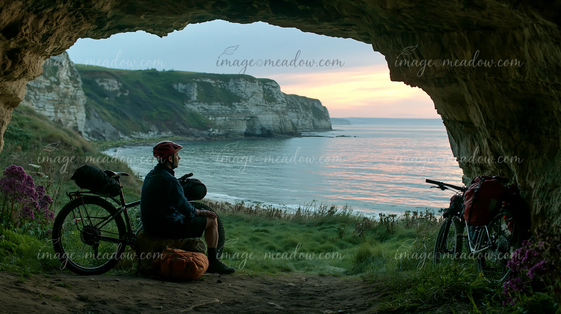 A cyclist rests inside a sea cave, overlooking the coastline at sunset.